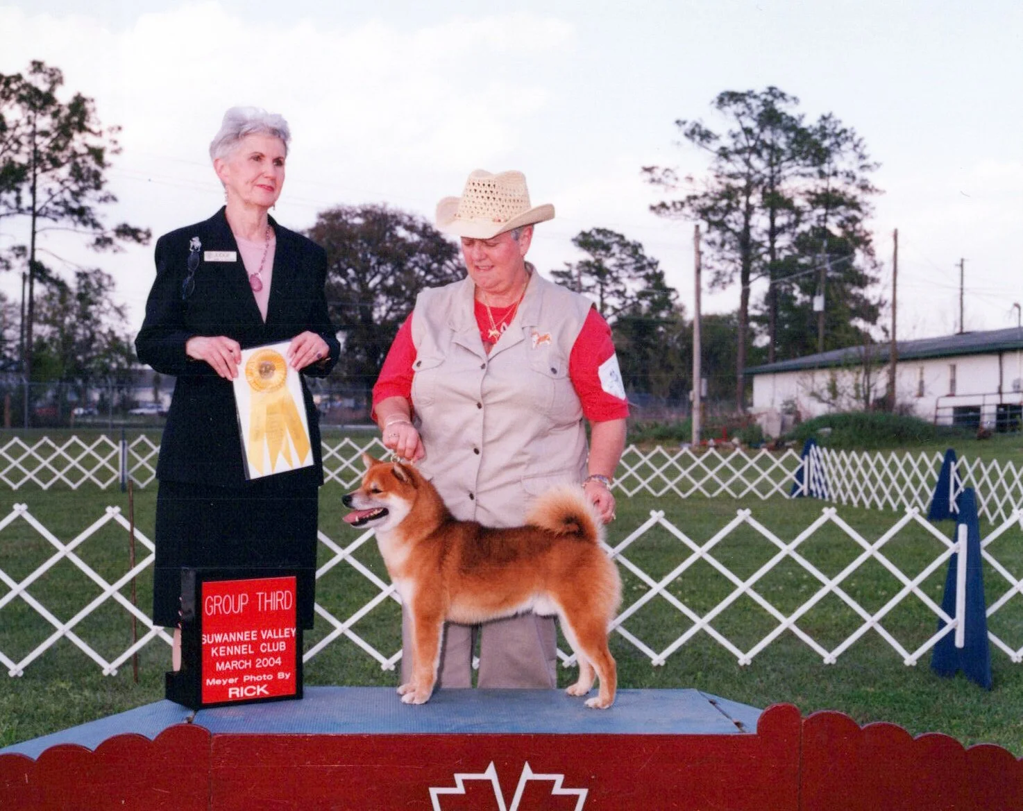 A woman and a dog at an outdoor dog show. The woman is wearing a cowboy hat and a beige vest, holding the dog on a platform. The woman beside her is dressed in a black suit and holding a yellow ribbon, standing next to a sign that reads 'Group Third,