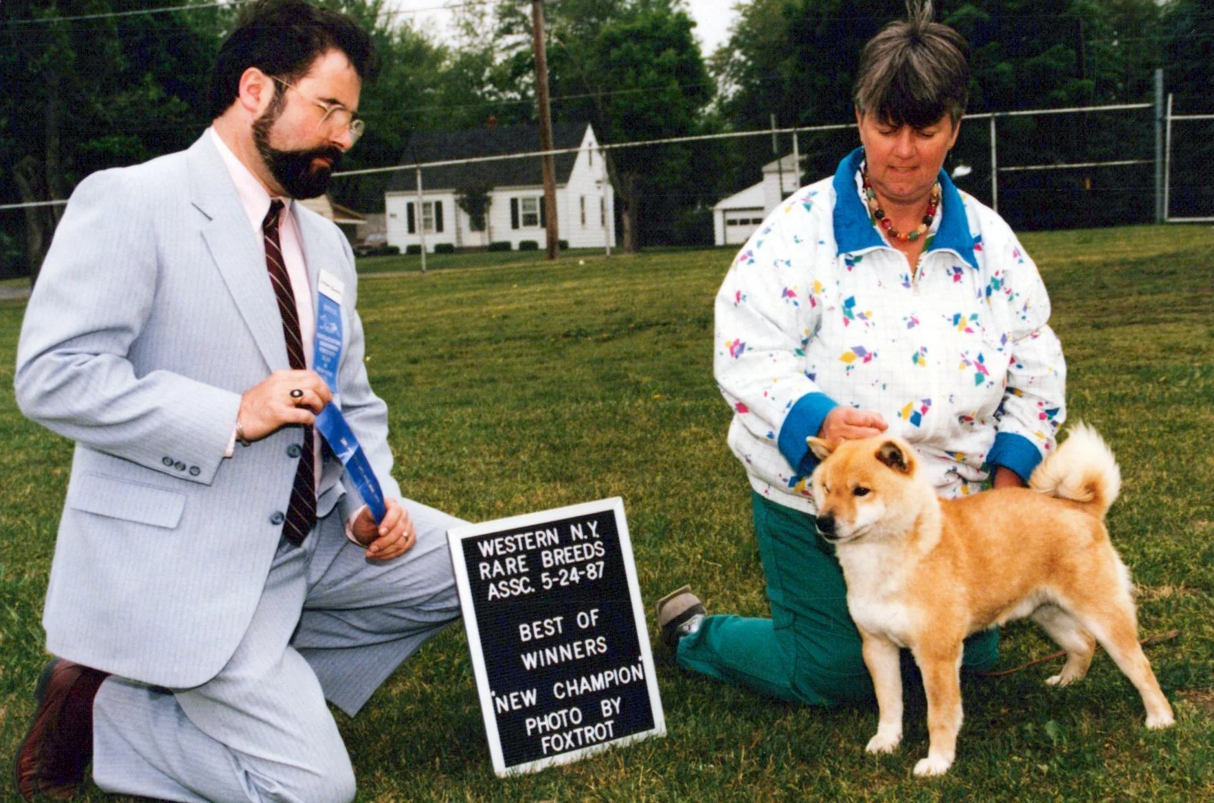 A man in a light gray pinstripe suit and a woman in a colorful jacket handling a Shiba Inu dog outdoors during a dog show. The man is kneeling with a blue ribbon, and the woman is petting the dog. A sign reads "Western N.Y. Rare Breeds, Assc. 5-24-87, Best of Winners, New Champion, Photo by Foxtrot." There are houses and trees in the background.