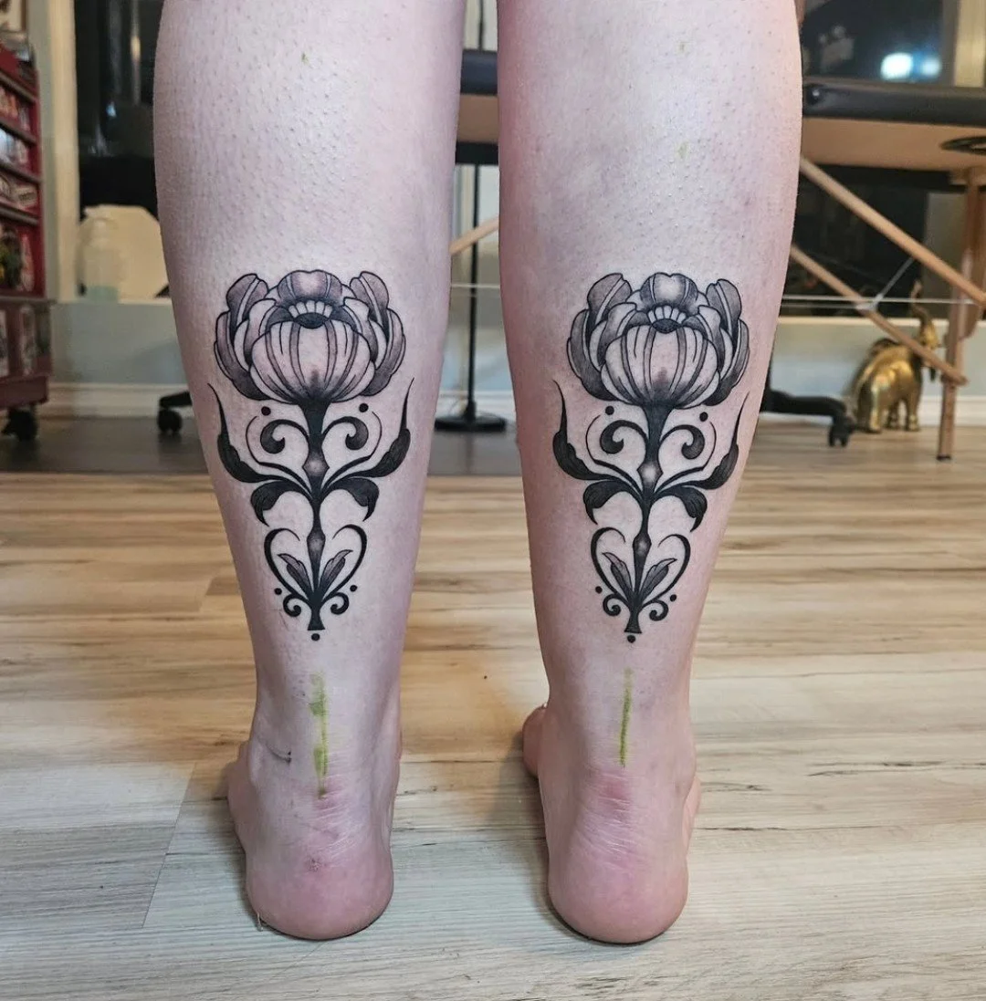 Tattoo of a symmetrical floral design on the back of each calf, featuring a large flower at the top with detailed petals and swirling patterns extending downward.