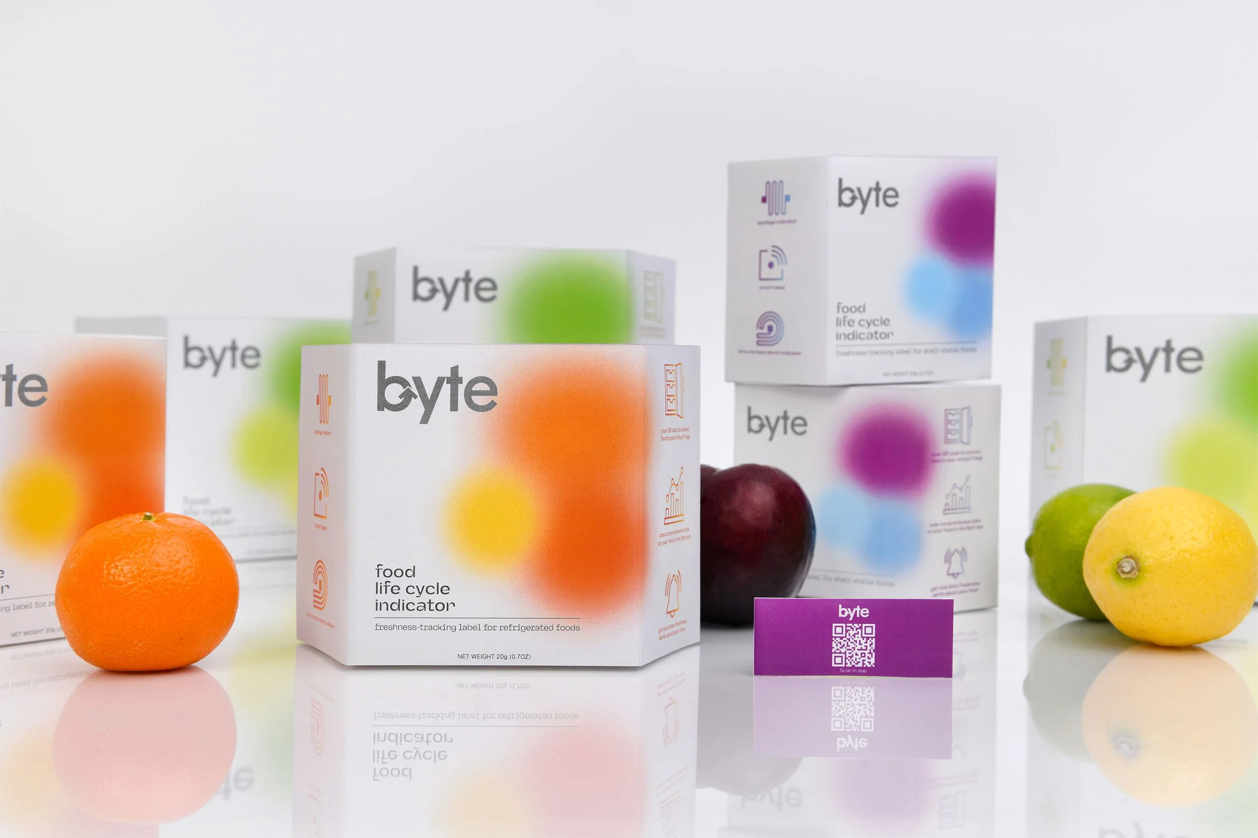Multiple packages of Byte food life cycle indicator boxes in front of fresh fruits including an orange, a plum, a lemon, and a lime, all on a reflective surface.