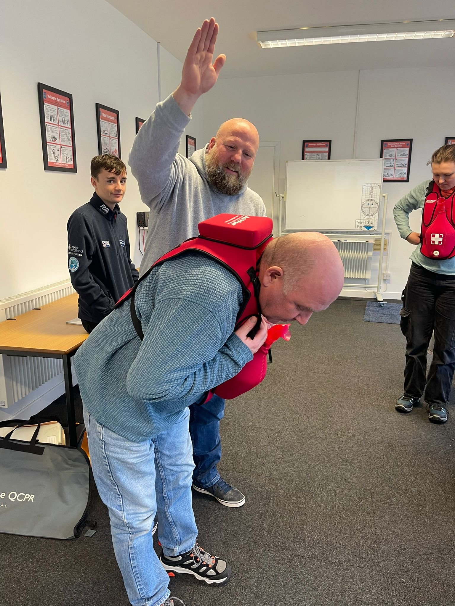 A man demonstrating CPR on a training dummy while another man watches and waves, with other people observing in a classroom setting.