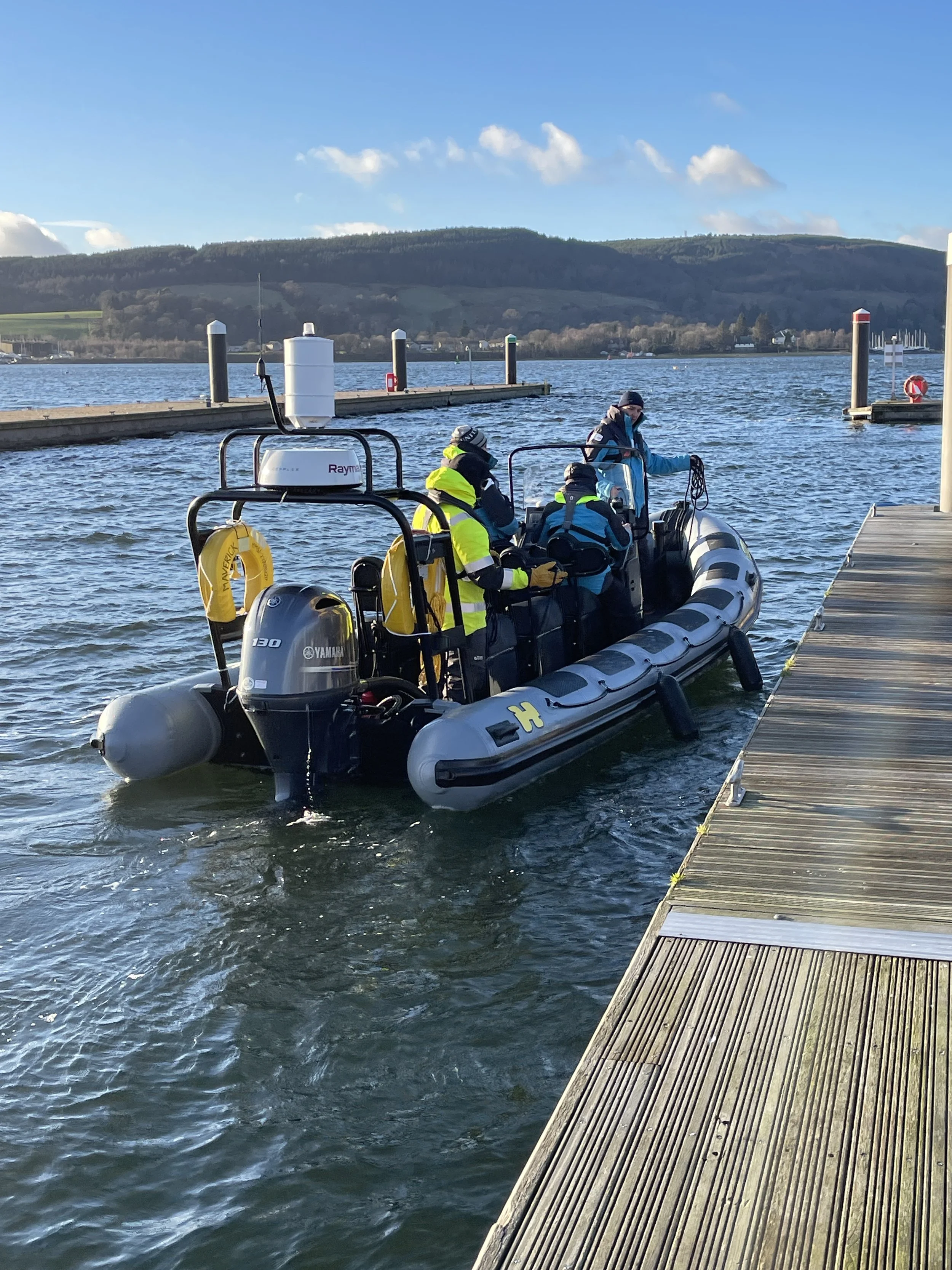 Students on an RYA Powerboat course learning to come alongside a wooden pier, with water, a mountain, and a partly cloudy sky in the background.