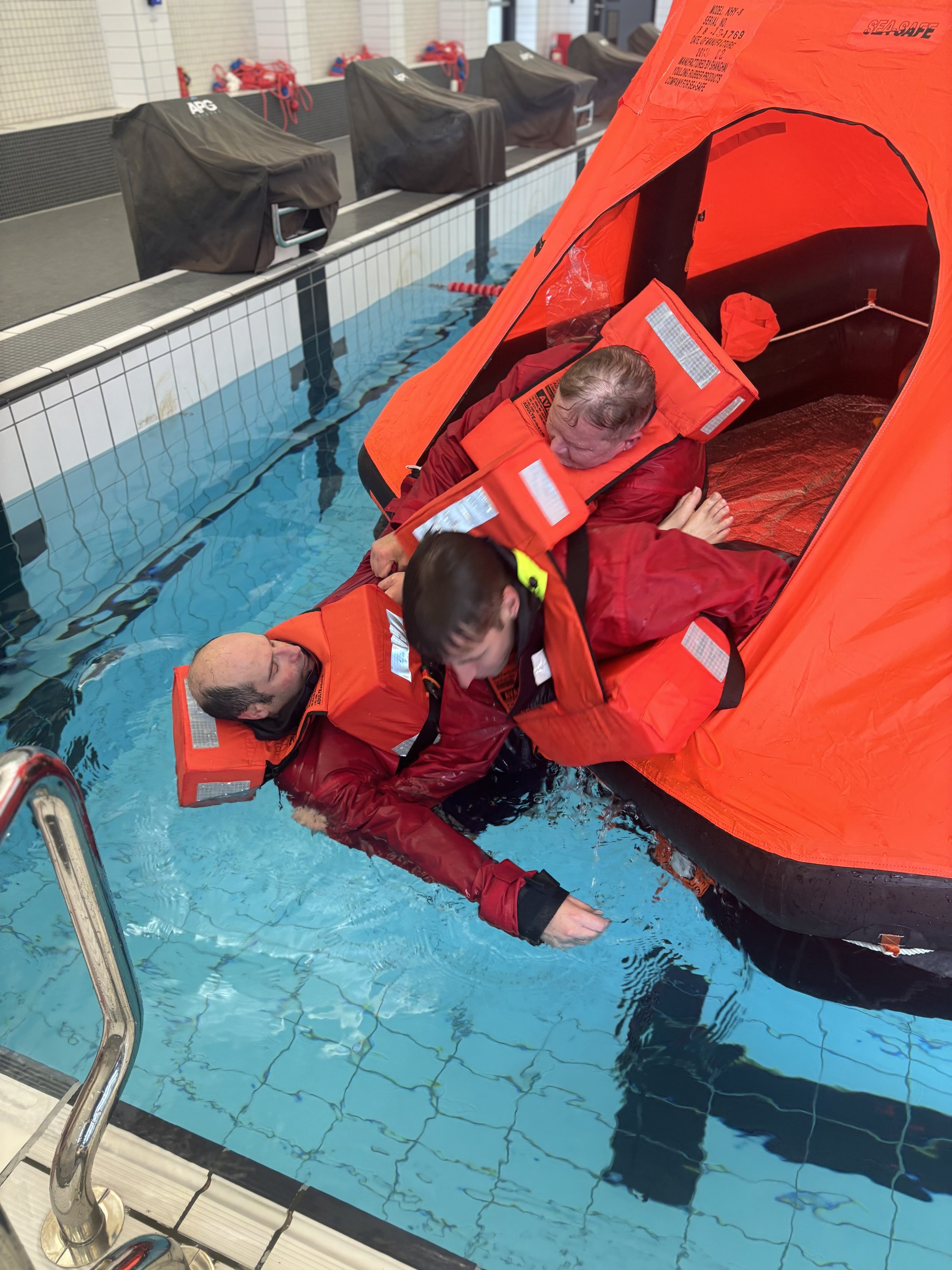 Rescue training exercise at an indoor swimming pool with individuals practicing assisting a person in the water to get into an inflated liferaft.