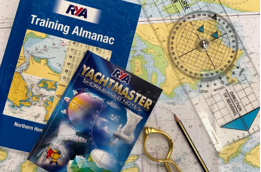Collection of nautical and navigation books and tools, including a Training Almanac, Yachtmaster Sharobed Notes, a compass, a nautical chart, a ruler, and a pencil.