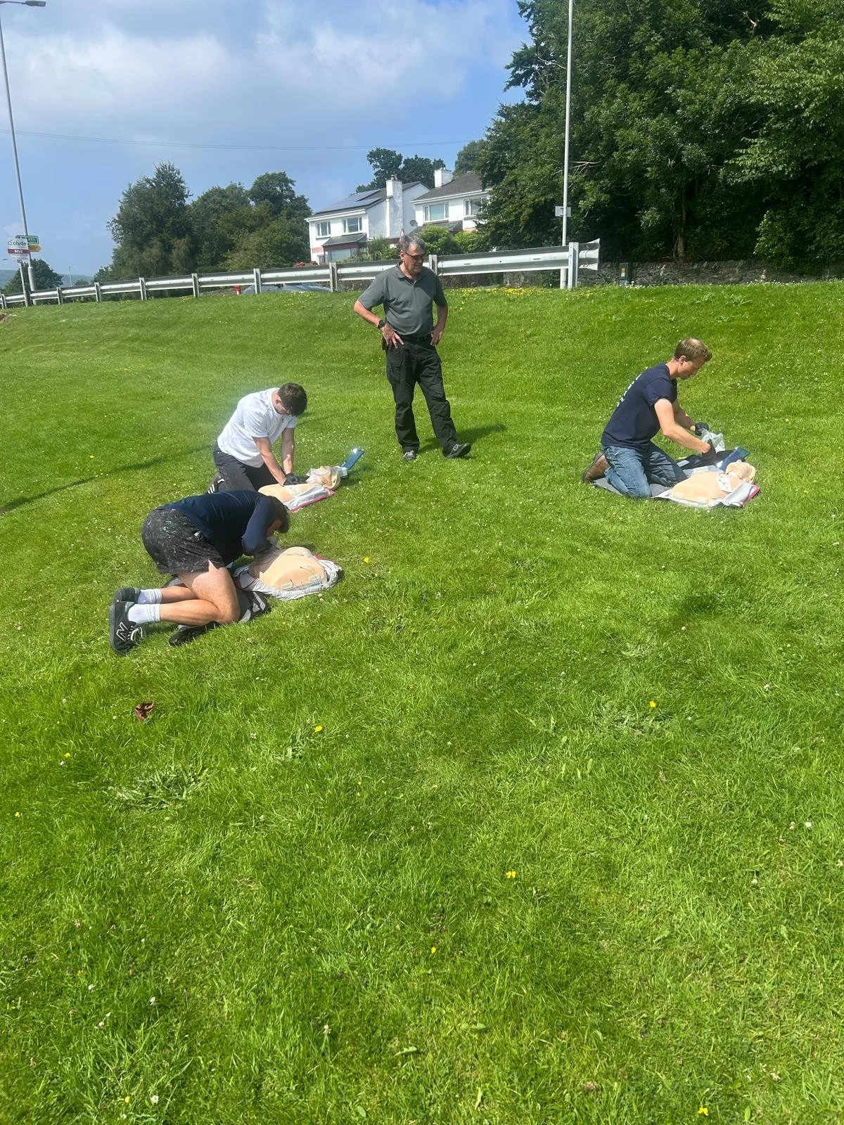 Four people practicing CPR on mannequins outdoors while an instructor observes.