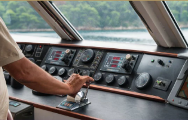 Close-up of a boat pilot's hand on the throttle control inside the boat's helm station, with navigation instruments and a window showing water and greenery outside.
