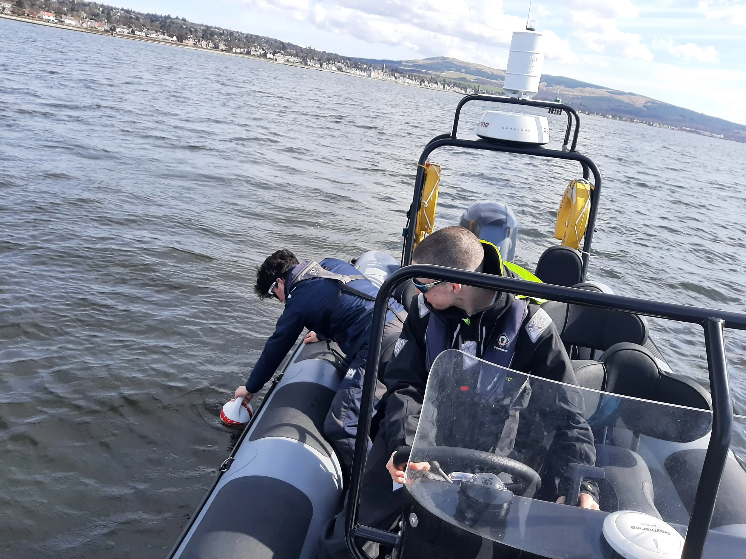Two people in a RIB, one leaning over the edge reaching into the water, and the other is sitting at the steering wheel, practicing man overboard procedure on an RYA Powerboat course