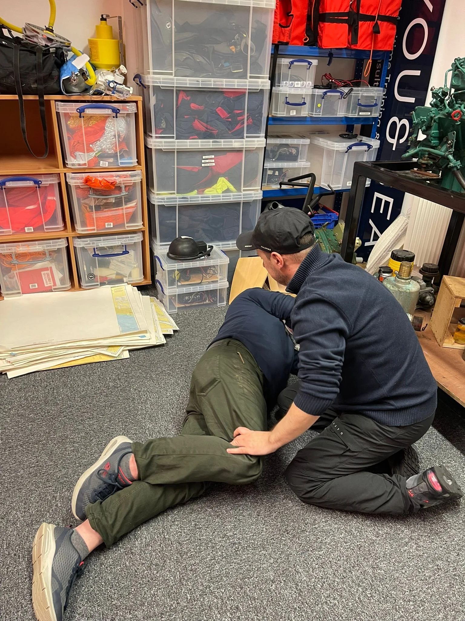 A person performing a first aid or CPR training on another person who is lying on the floor. The setting appears to be a storage room or workshop with plastic bins, shelves, and equipment around.
