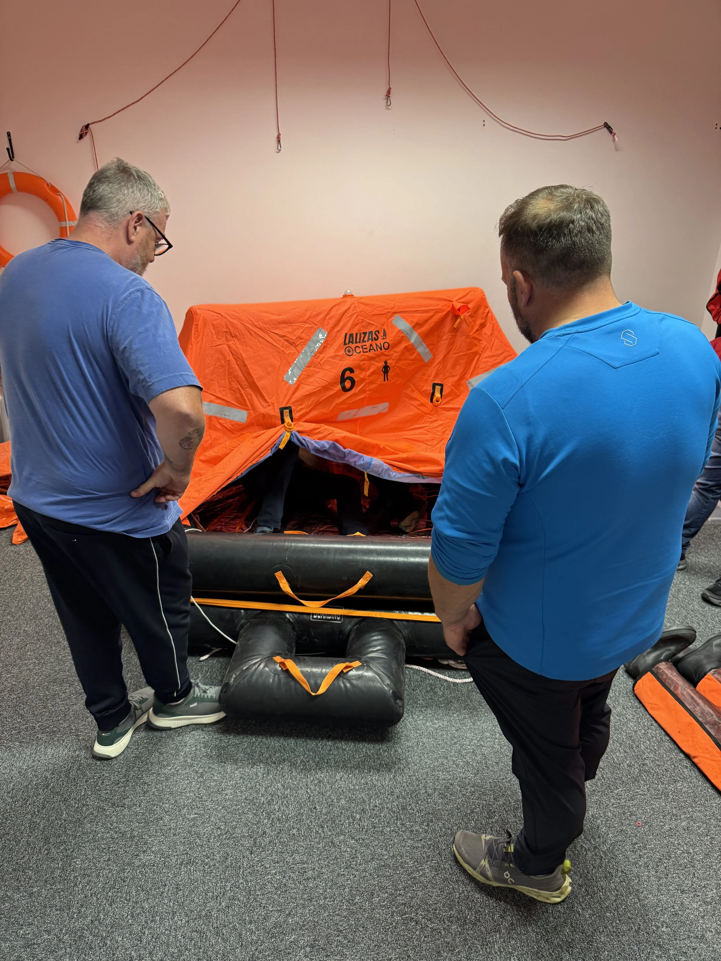 Two men standing next to an inflated liferaft with a person inside, in an indoor setting.  The men are observing or preparing the rescue equipment.