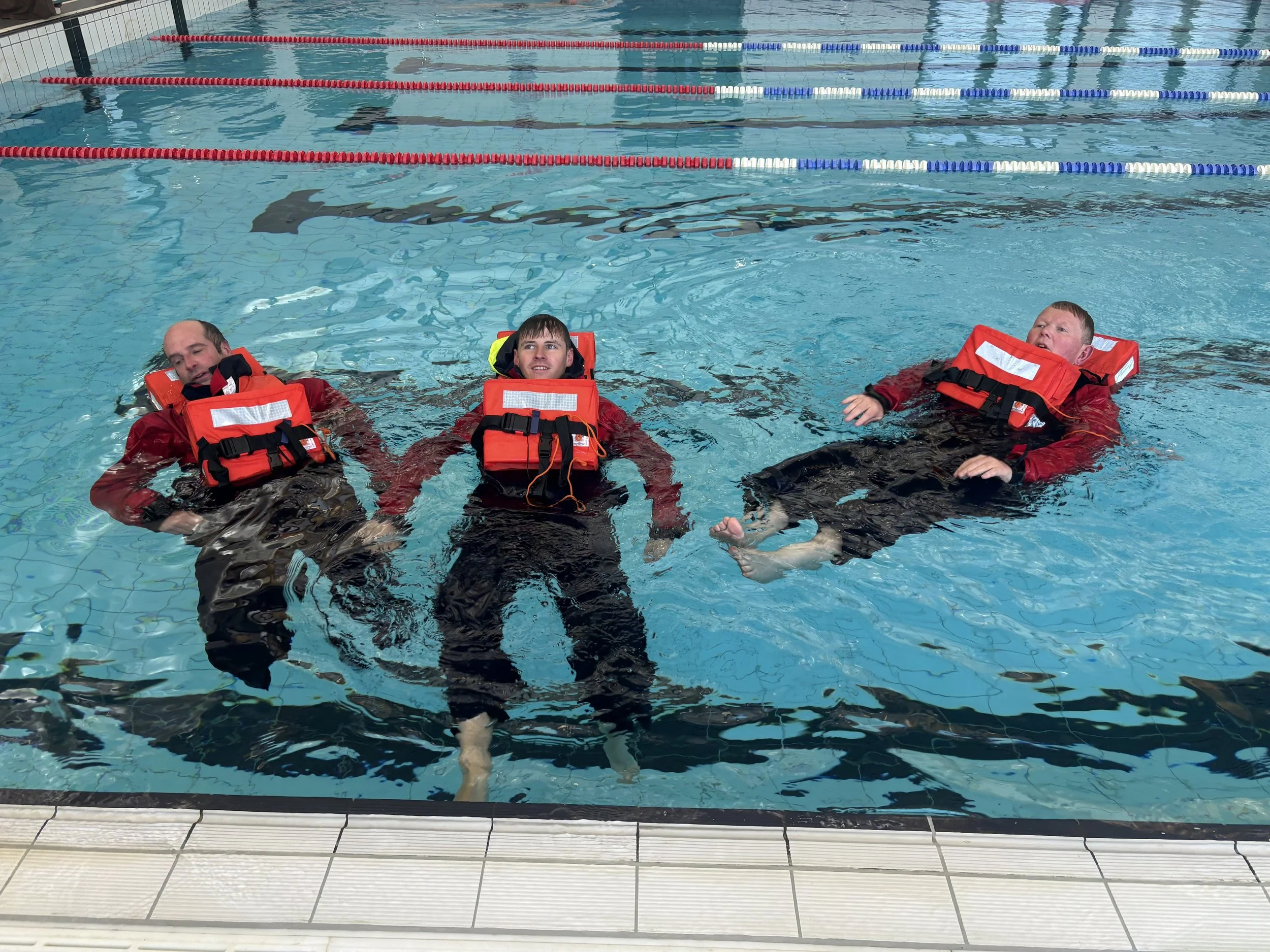 Three people floating on their backs in a swimming pool wearing red life jackets.