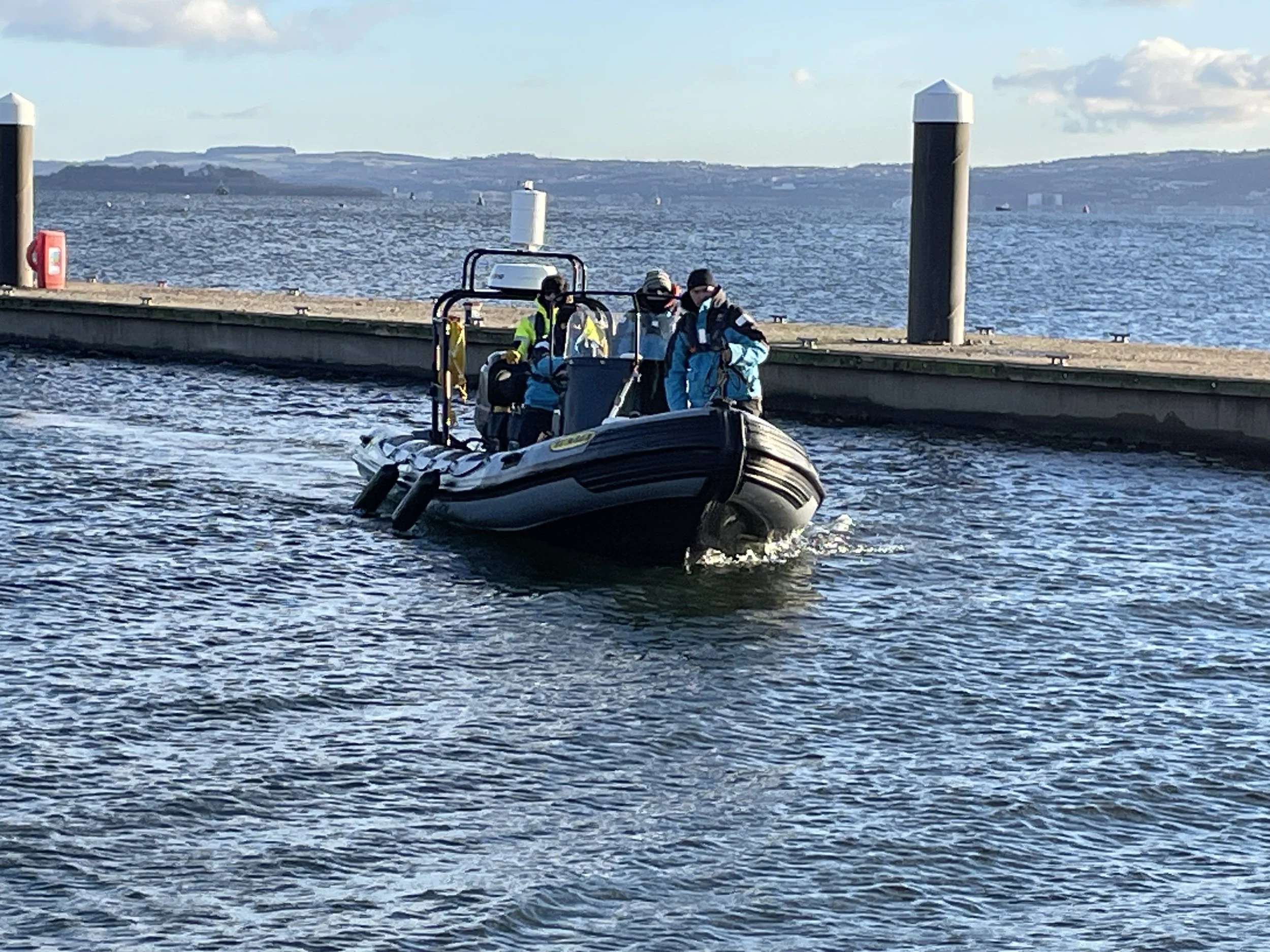 students on a rIB during an RYA Powerboat course, all wearing jackets and safety gear, approaching a pontoon, under a partly cloudy sky.