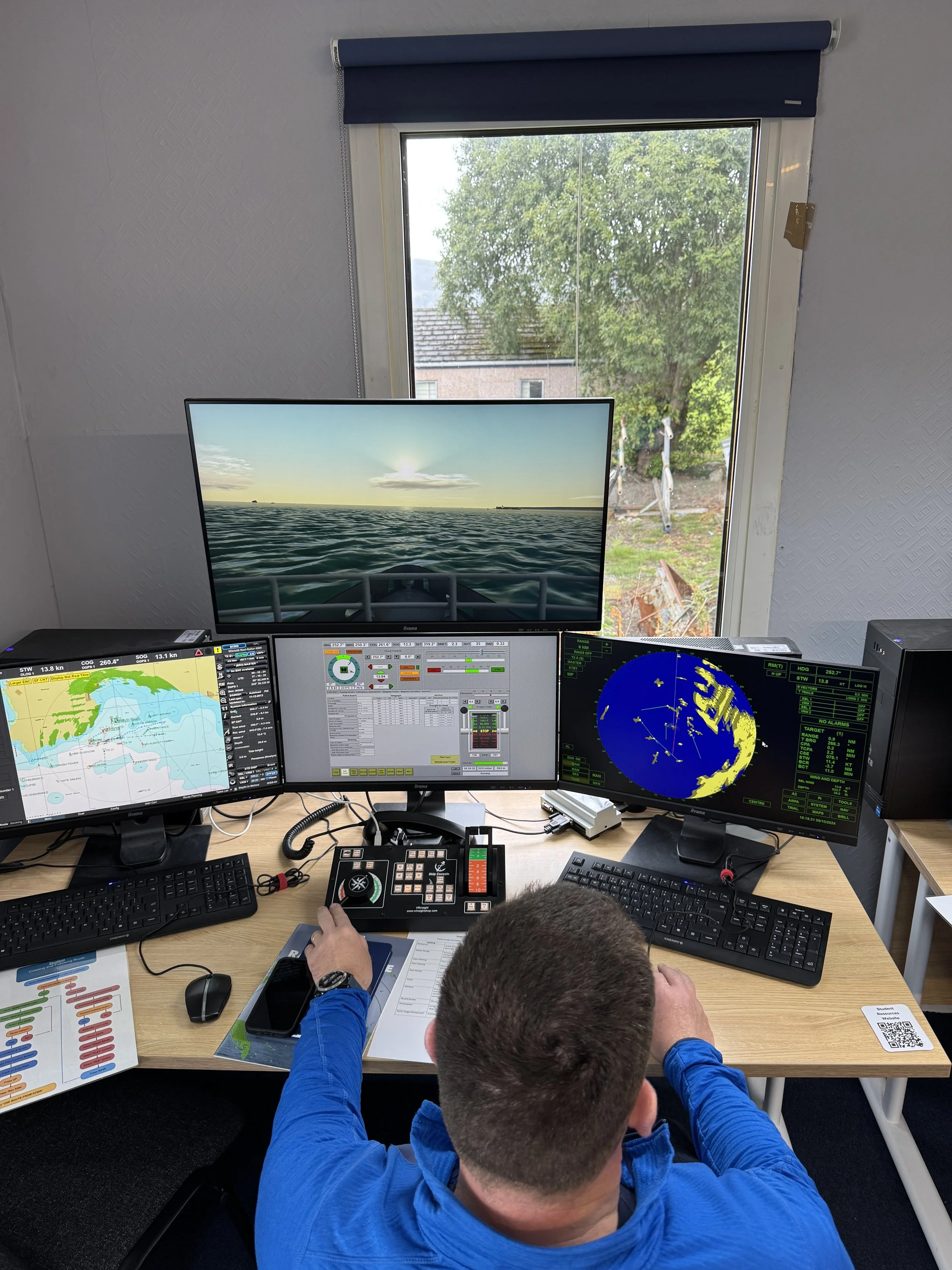 A student using the Transas simulator's multiple computer monitors displaying navigation and radar screens in a ship's bridge setup.