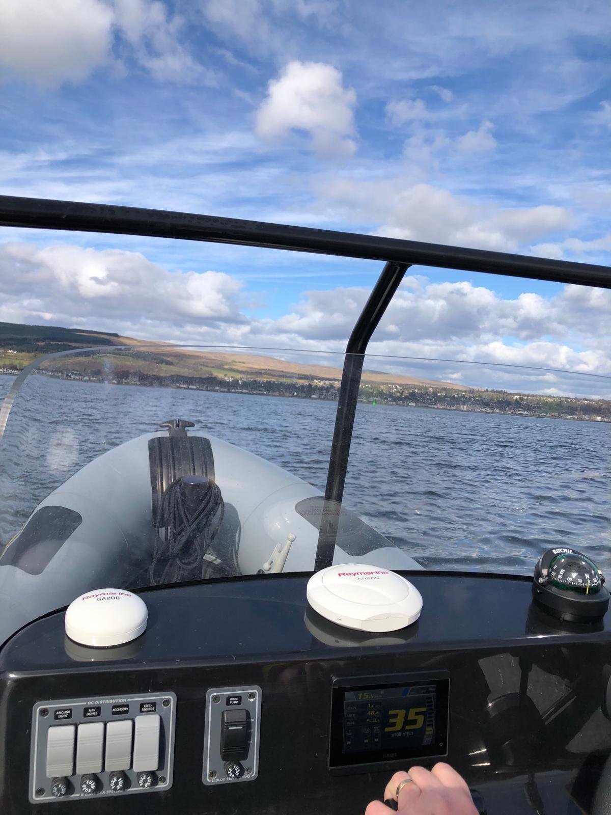 View from a boat dashboard showing a digital screen, two Raymarine devices, and a green compass. The boat is on the water with land and hills in the distance, under a partly cloudy sky.