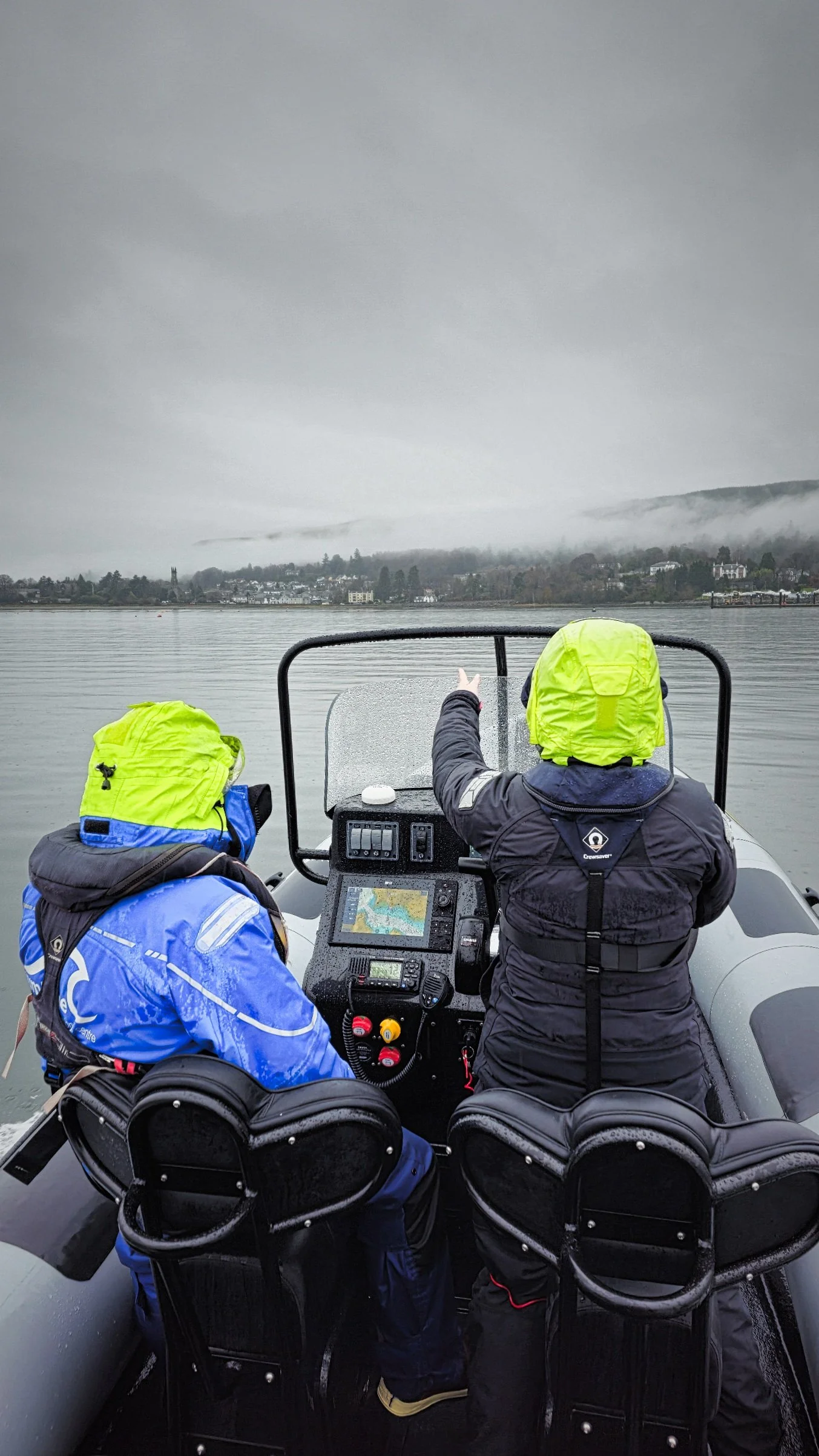 Two students on an RYA Powerboat course, wearing rain gear, in a RIB, with one pointing at a misty, cloudy shoreline across a calm body of water.