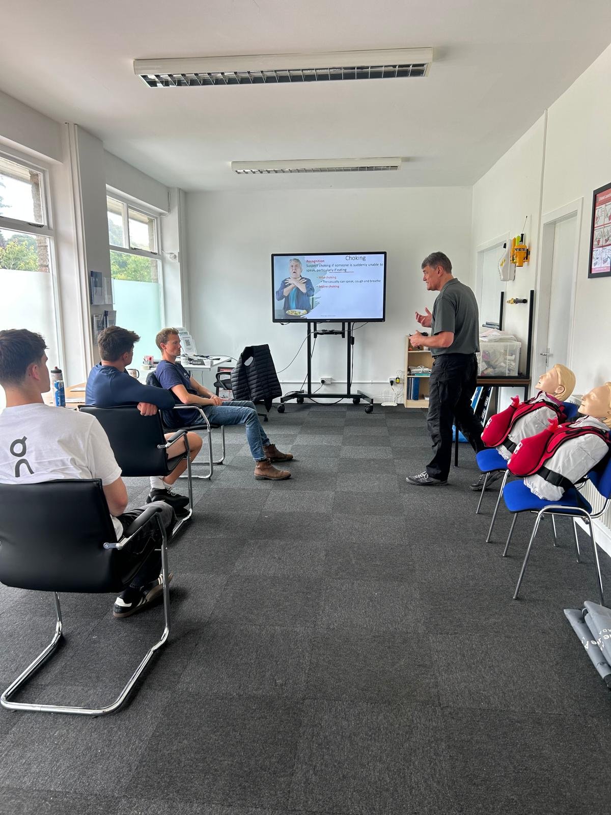 A instructor is giving a CPR training class to a group of young men in a classroom. The instructor is standing next to a large screen displaying a presentation on choking and first aid. The students are seated facing the instructor, with medical mannequins in backpacks on the side.