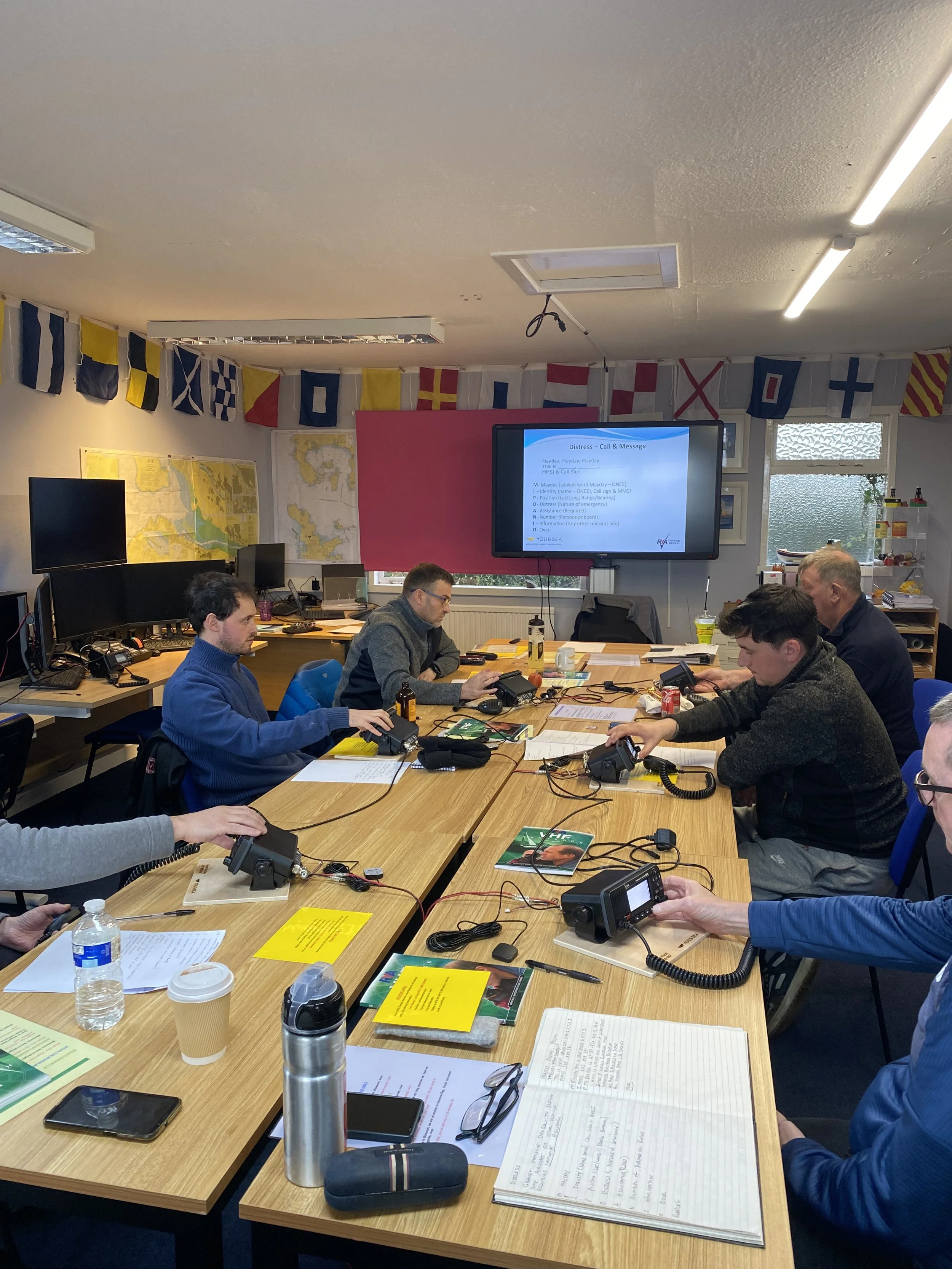 Students in a classroom, with a large screen displaying a presentation titled 'Distress - Call & Message', on the RYA VHF Radio course