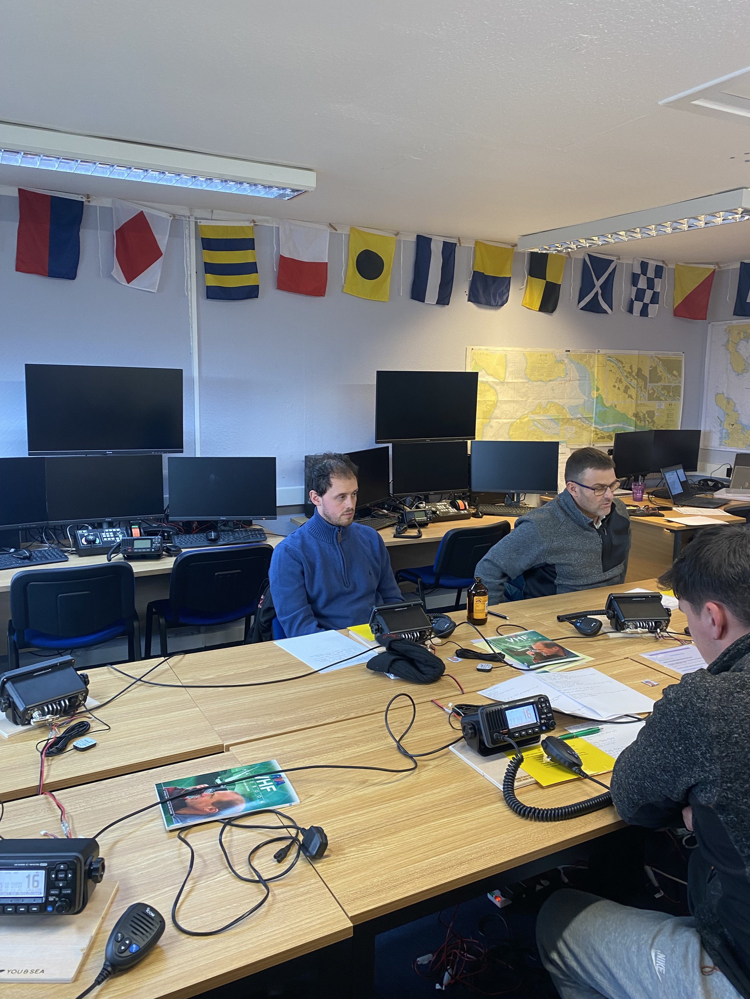 Students sitting around a large table in a conference room with multiple computer monitors, flags hanging on the wall, and a map in the background, on one of You & Sea's maritime training courses