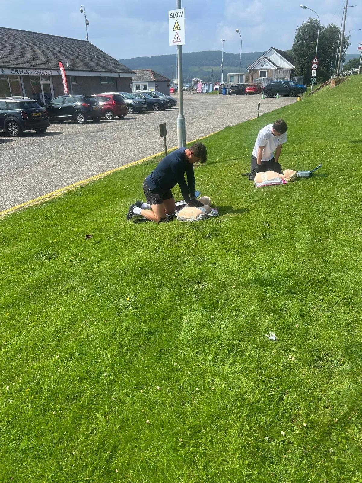 Two individuals performing CPR on mannequins outdoors on a grassy area with parking lot and buildings in the background.