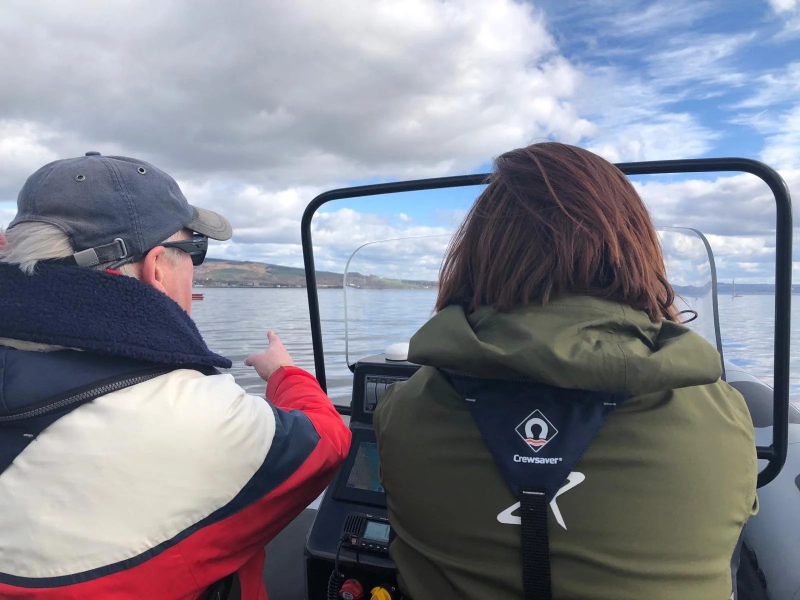 In a rIB on the water, an RYA instructor advises a student, looking at a navigation device, while pointing ahead. The RIB is on a large body of water with a shoreline and hills in the distance, under a partly cloudy sky.
