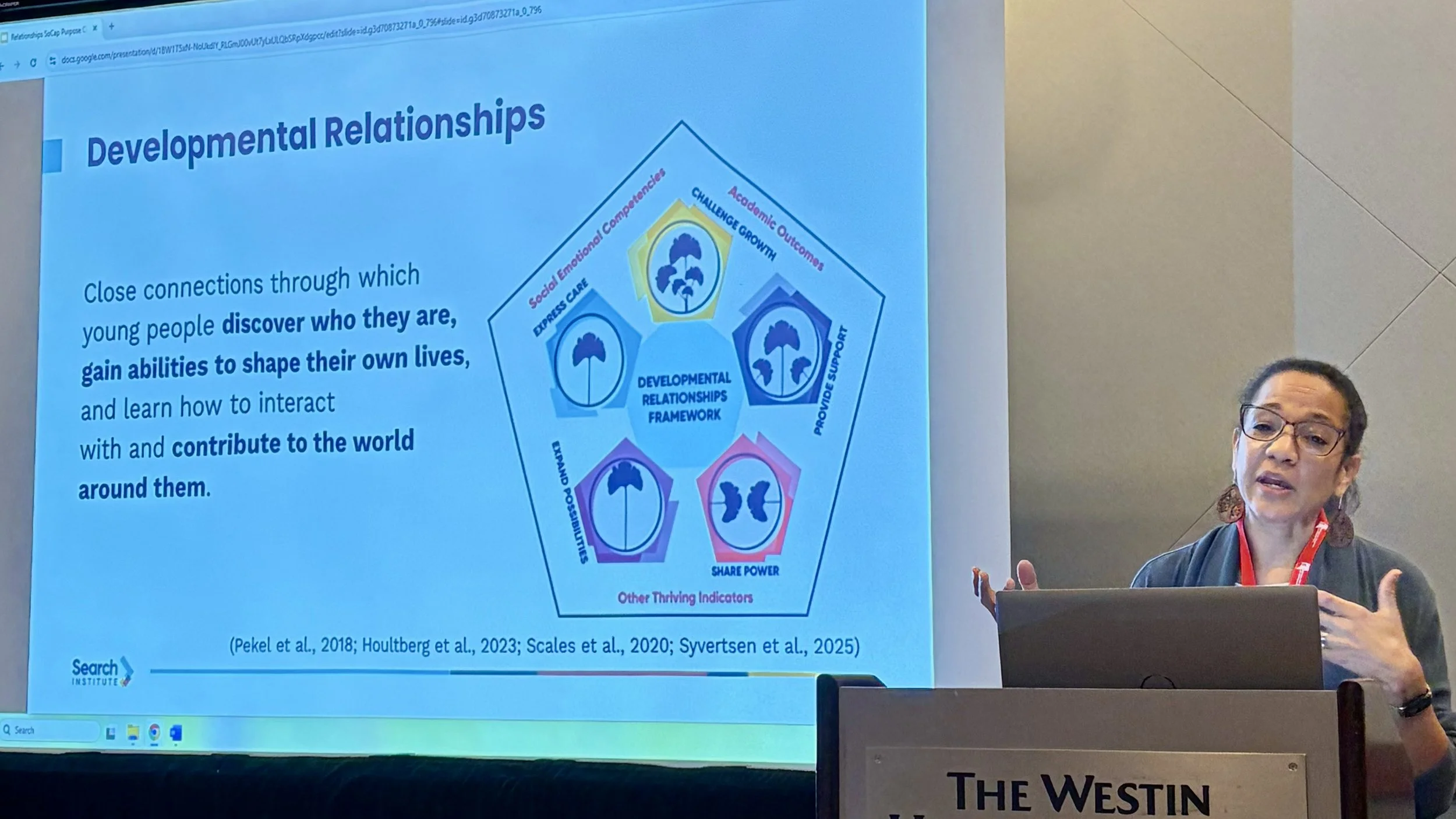 Joanna Williams, from the Search Institute, presents research during a conference. Williams is standing at dais with a screen to her right.