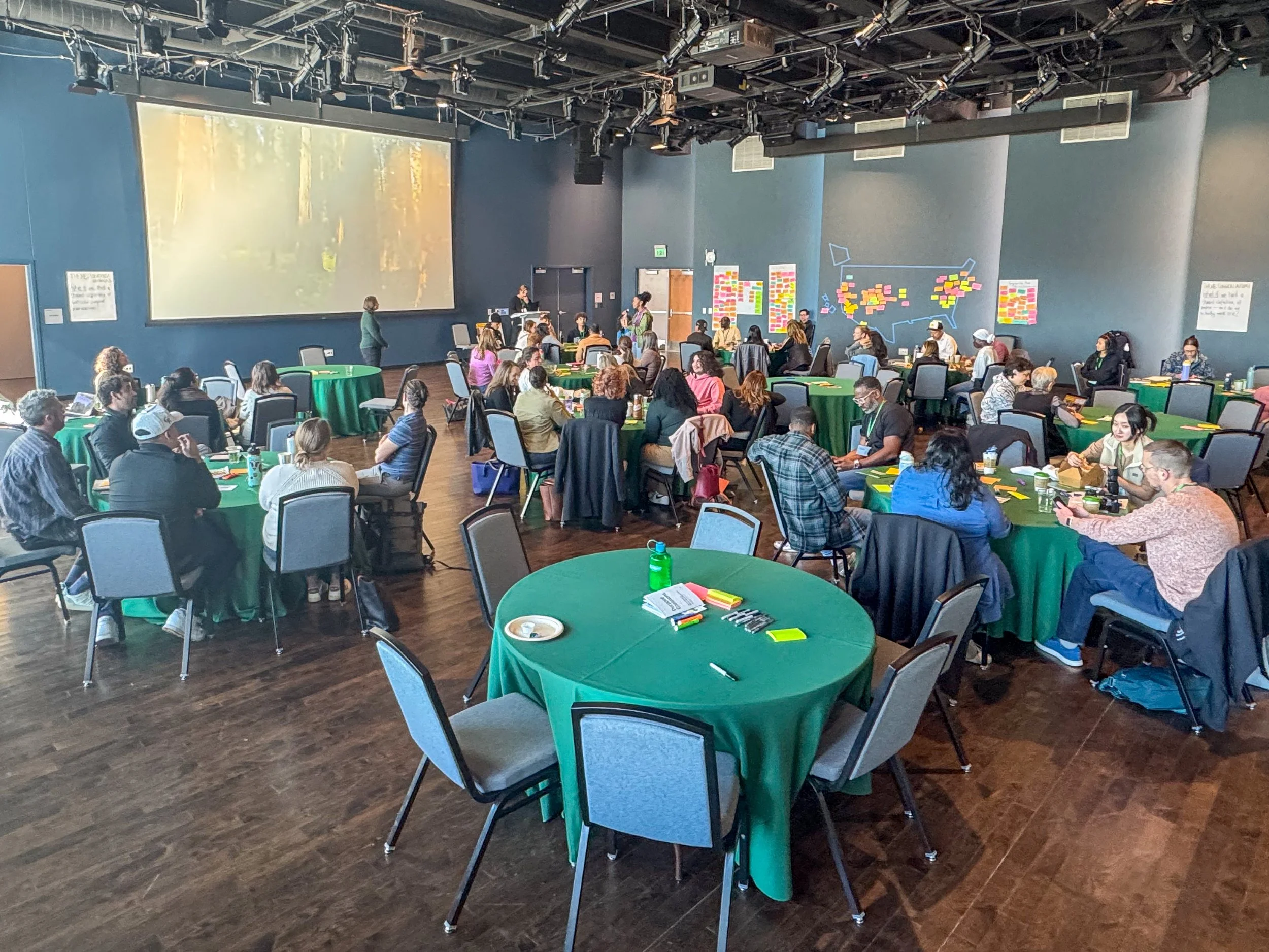 A photo of groups of people, sitting at tables, working together on a project.