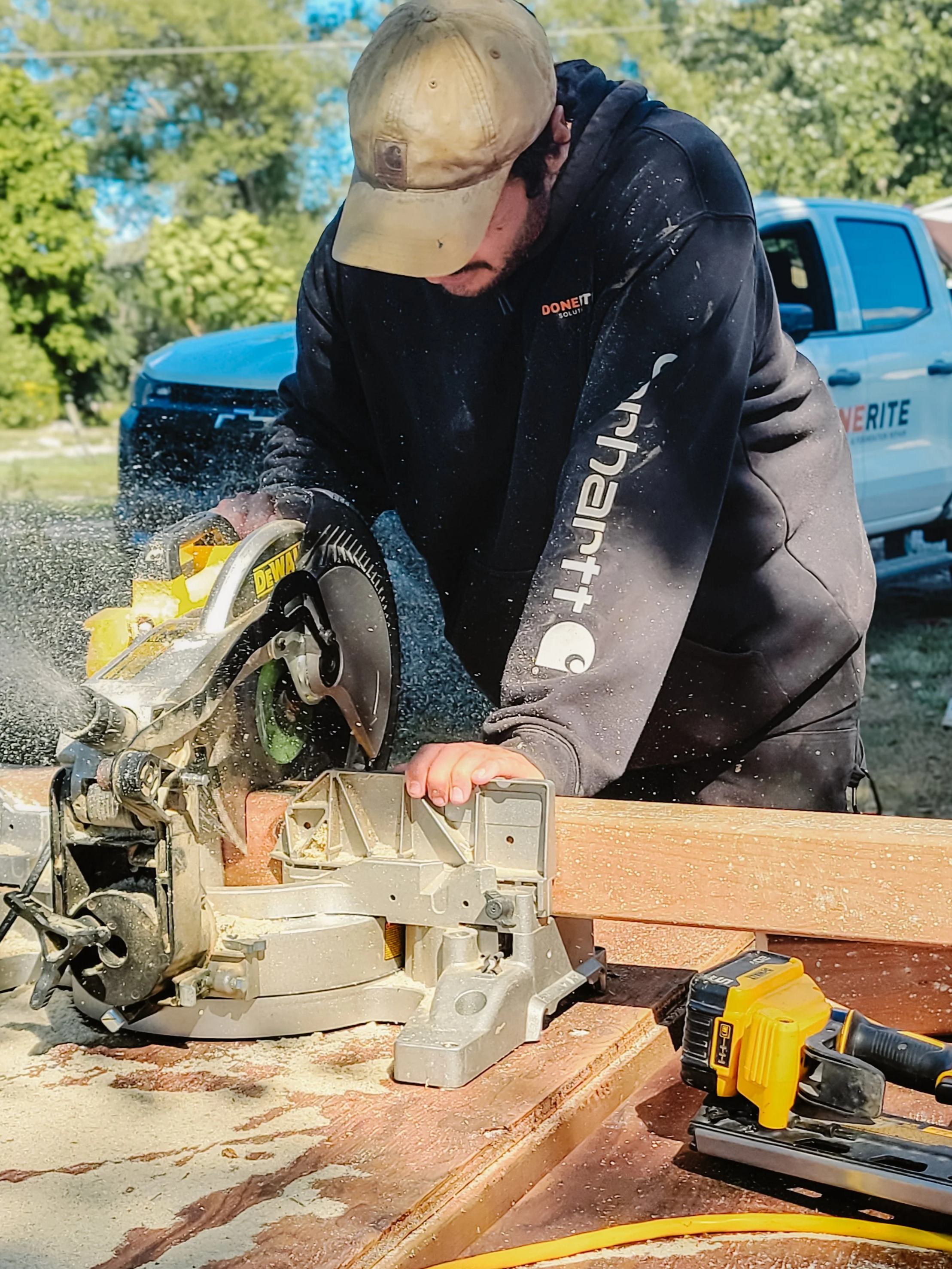 A man operating a circular saw outdoors, cutting a piece of wood on a workbench, with a pickup truck and green trees in the background.
