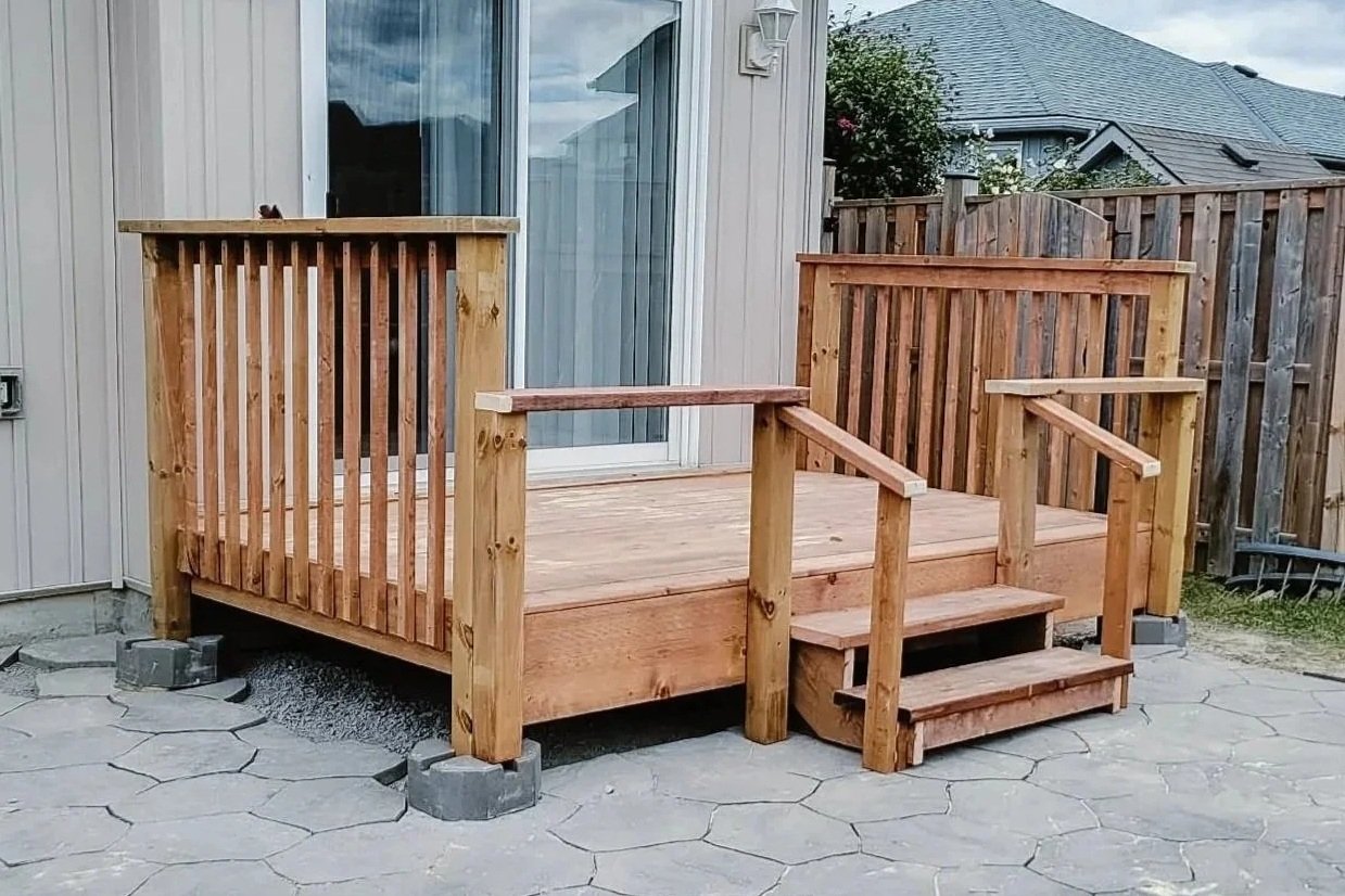Wooden deck with stairs and railing attached to the back of a house, with a sliding glass door, on a stone patio, surrounded by a wooden fence and neighboring houses.