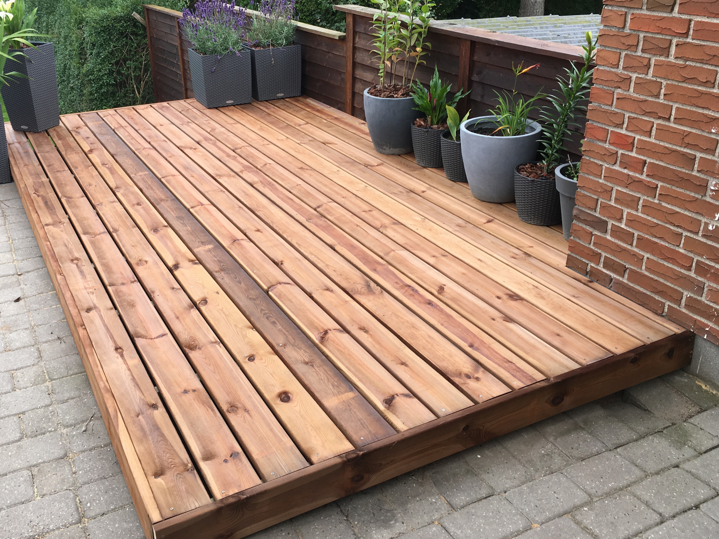 A wooden deck outdoor area with potted plants and a brick wall.