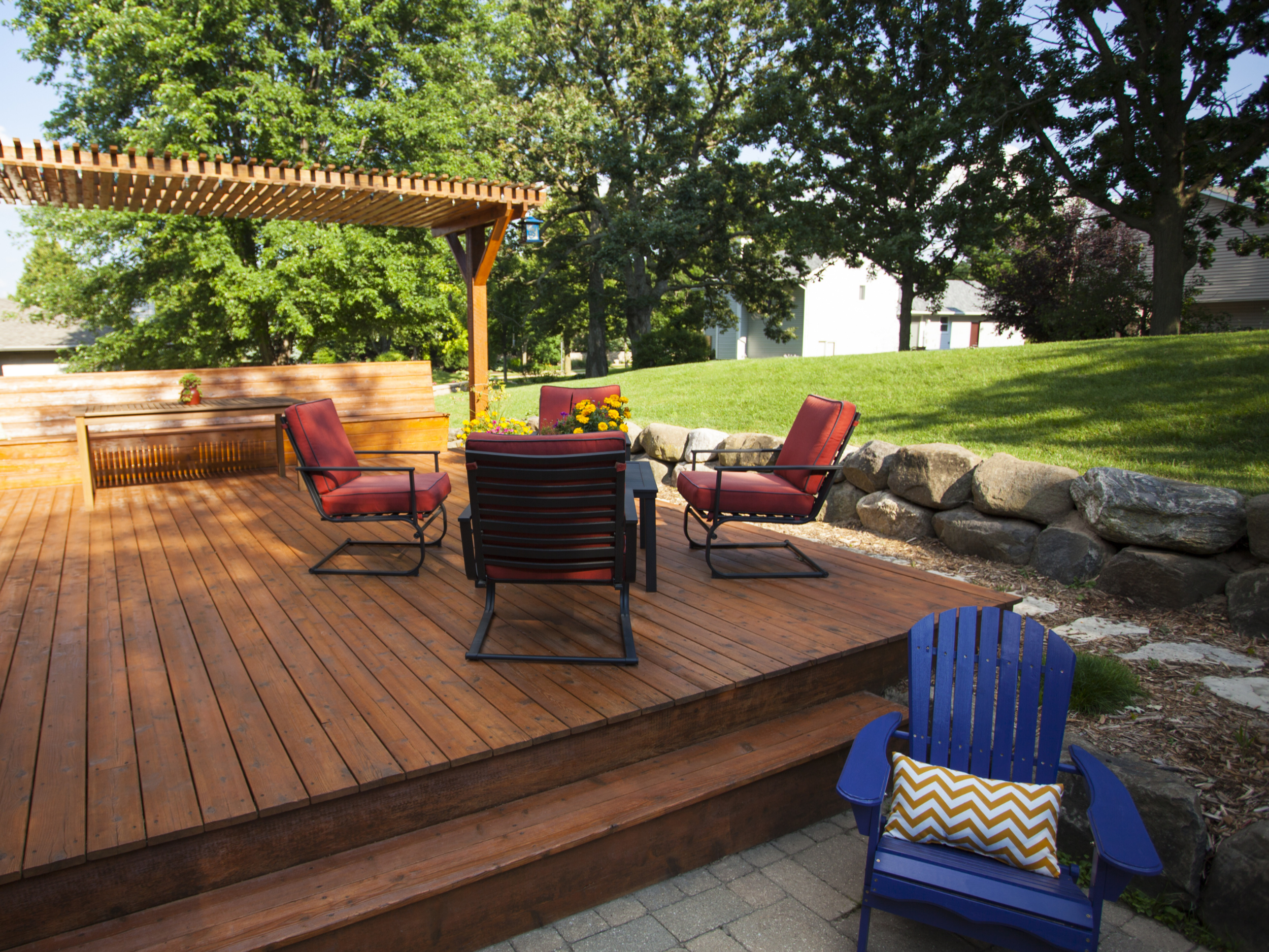 A backyard patio with a wooden deck, outdoor chairs with red cushions, a blue Adirondack chair with a chevron pillow, a stone border, green grass, trees, and a house in the background.