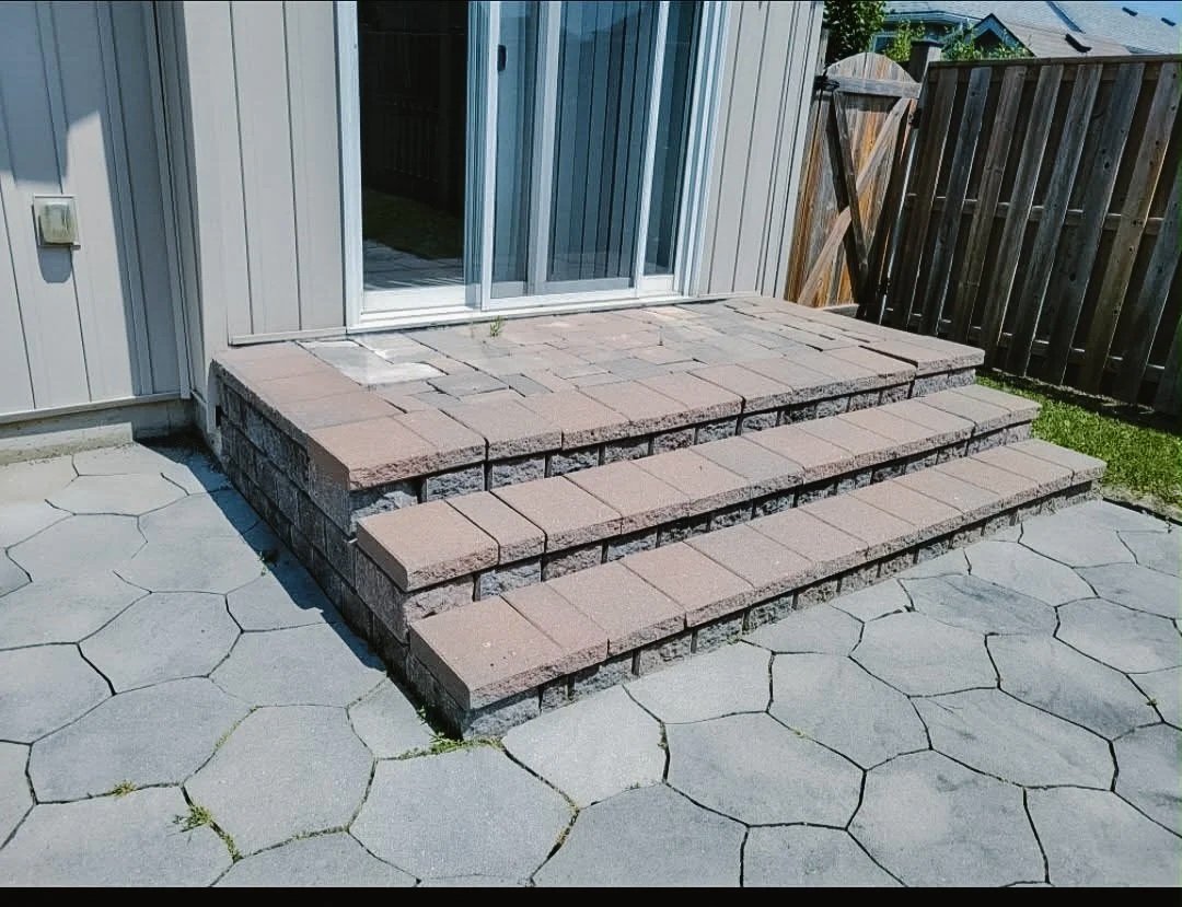 A set of three brick steps leading up to a sliding glass door on a house with white siding, surrounded by a concrete patio and a wooden privacy fence.