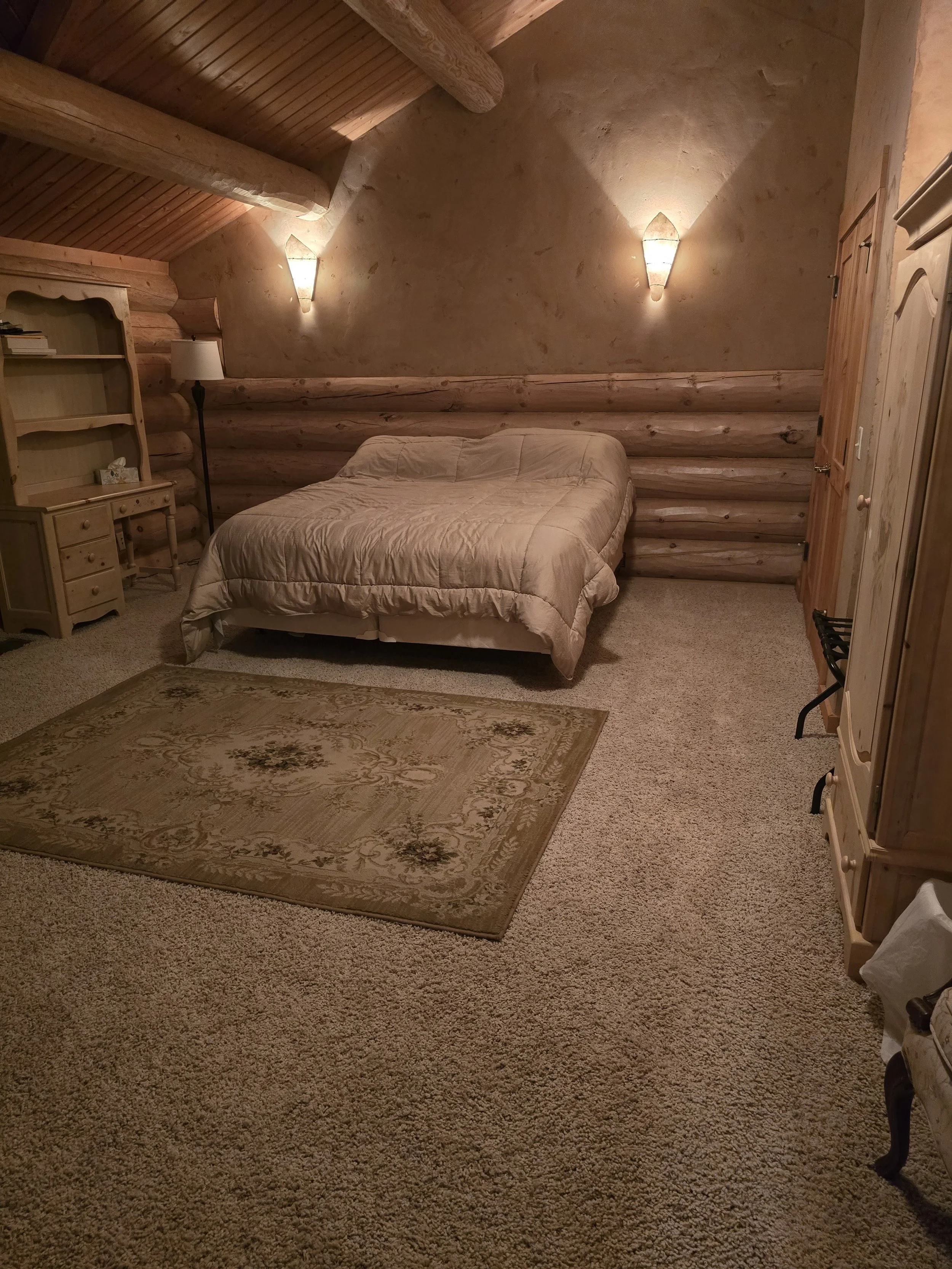 Cozy bedroom with a rustic log cabin design, featuring a wooden bed, soft beige bedding, carpeted floor, area rug, wooden furniture including a side table and cabinet, and soft lighting from wall sconces.