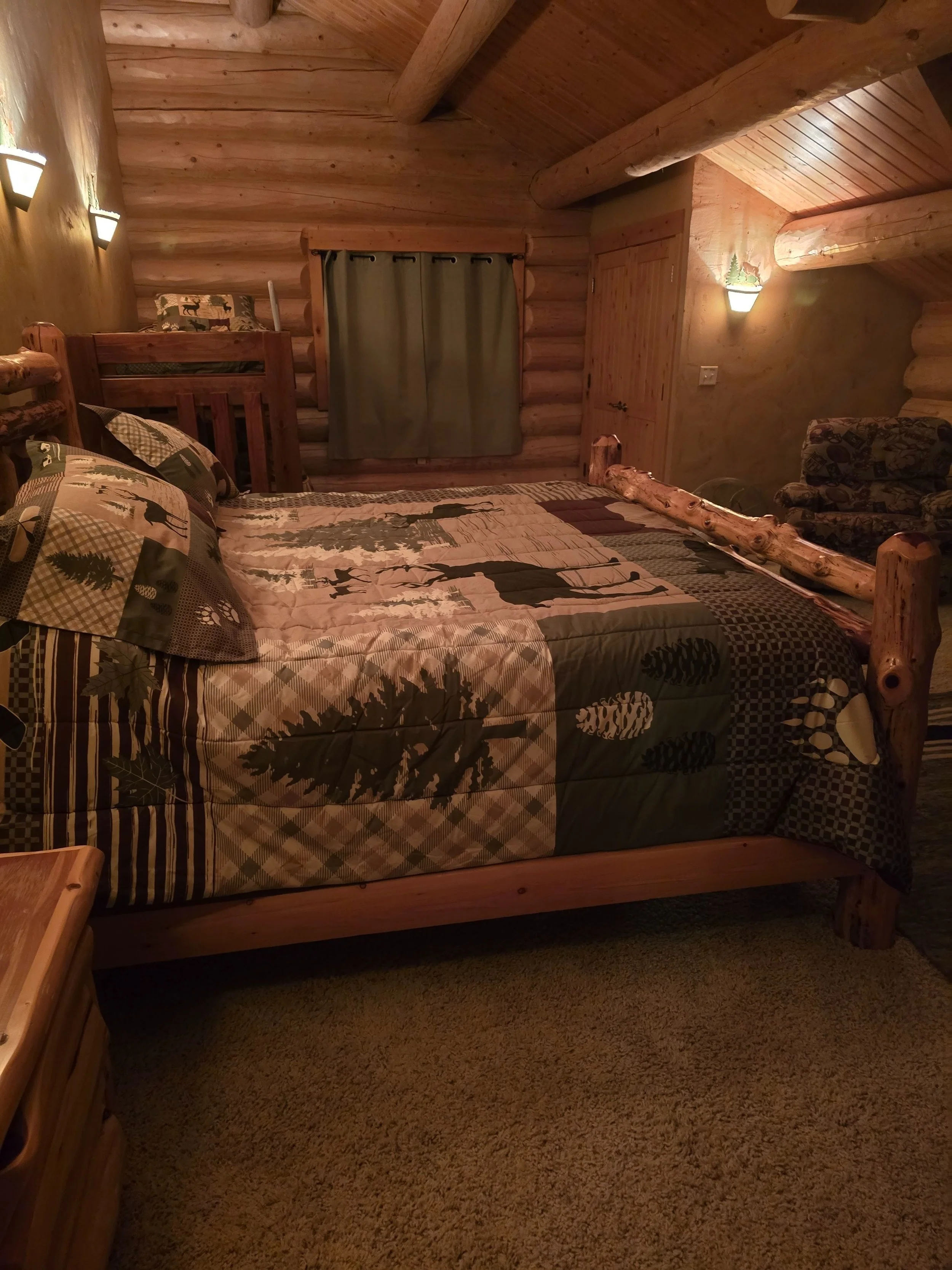 Rustic log cabin bedroom with a wooden bed frame, patterned quilt featuring nature motifs, log walls and ceiling, green curtains, wall sconces, and upholstered armchair.