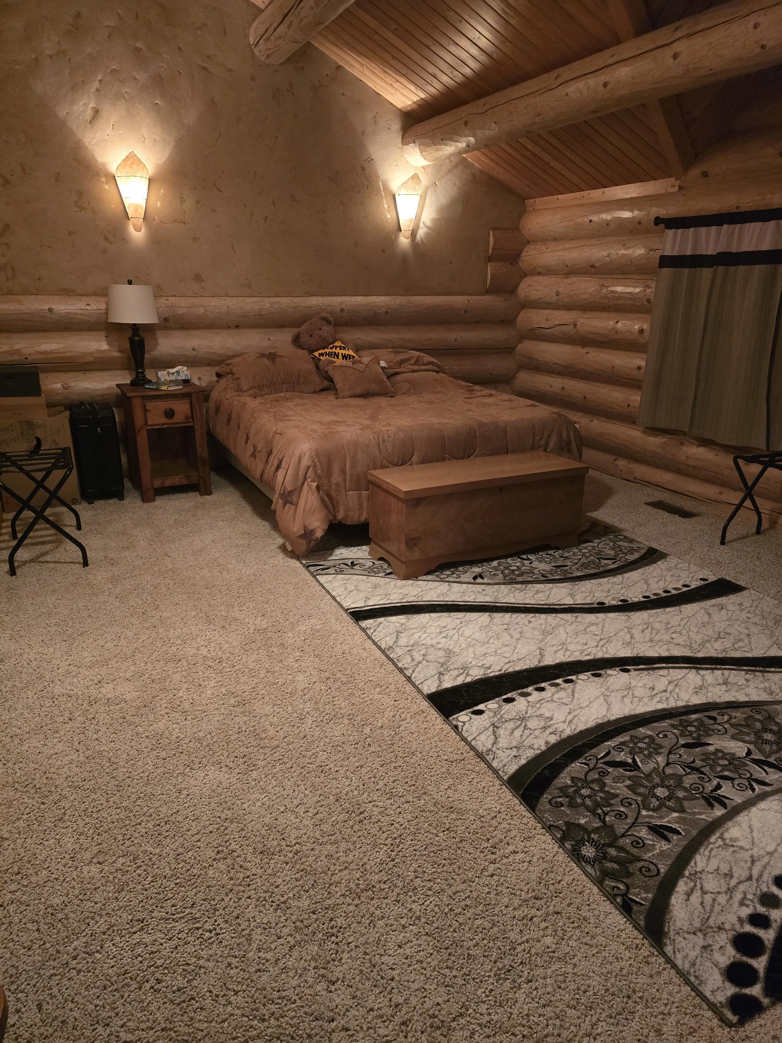 Cozy log cabin bedroom with wood-paneled walls and ceiling, featuring a bed with brown bedding, bedside table with lamp, patterned rug, and ambient lighting.