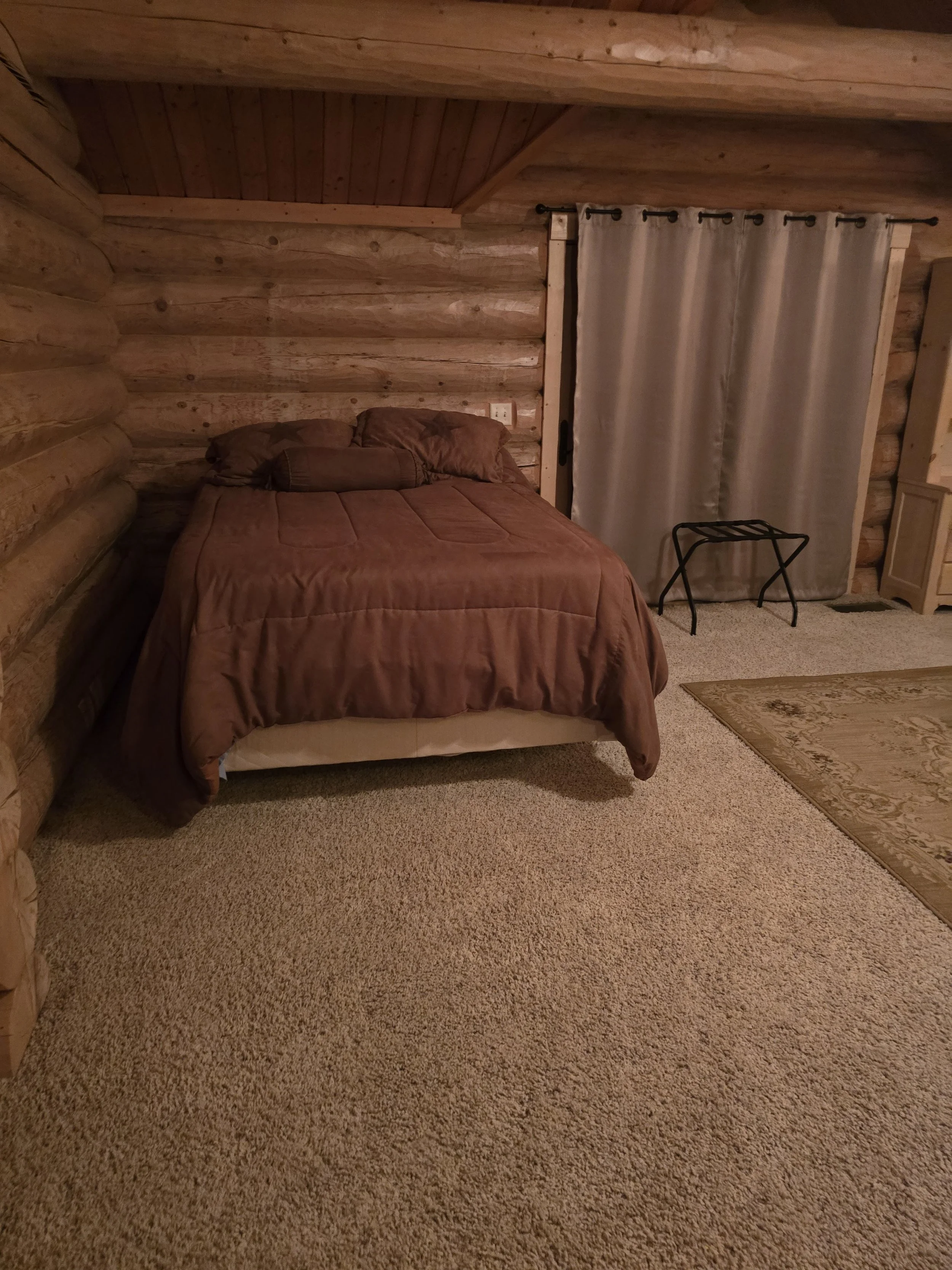 Cozy log cabin bedroom with wooden walls and ceiling. Features a bed with brown bedding, a side table with luggage rack, and carpeted floor.