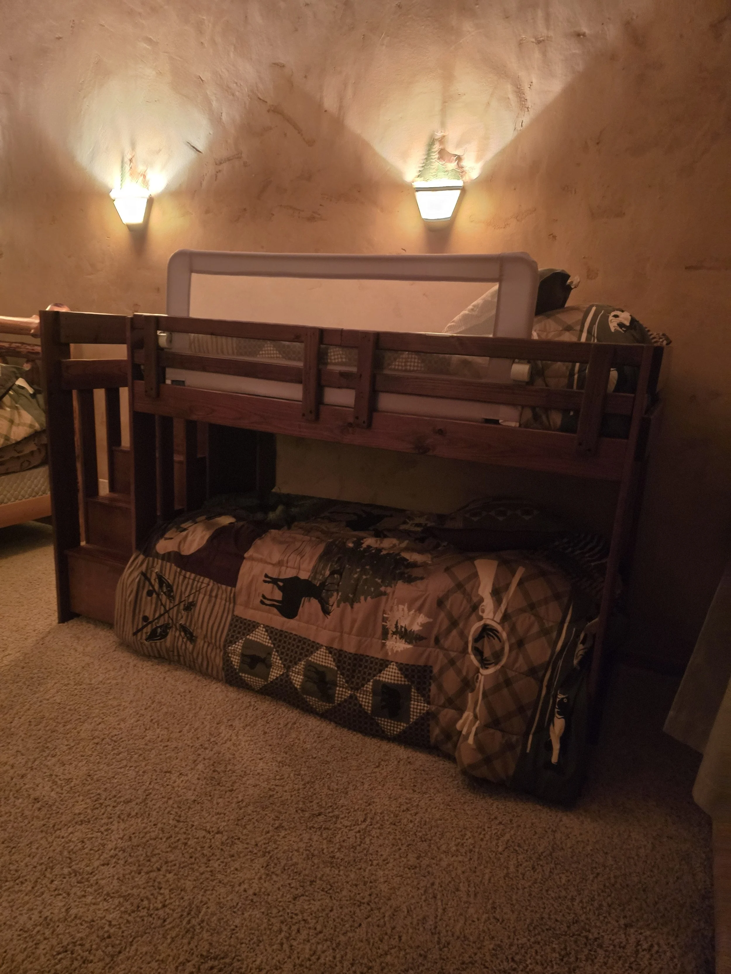 Bunk bed with rustic-themed bedding in a dimly lit room, featuring two wall-mounted lights, a safety rail on the upper bunk, and a wood-paneled wall.