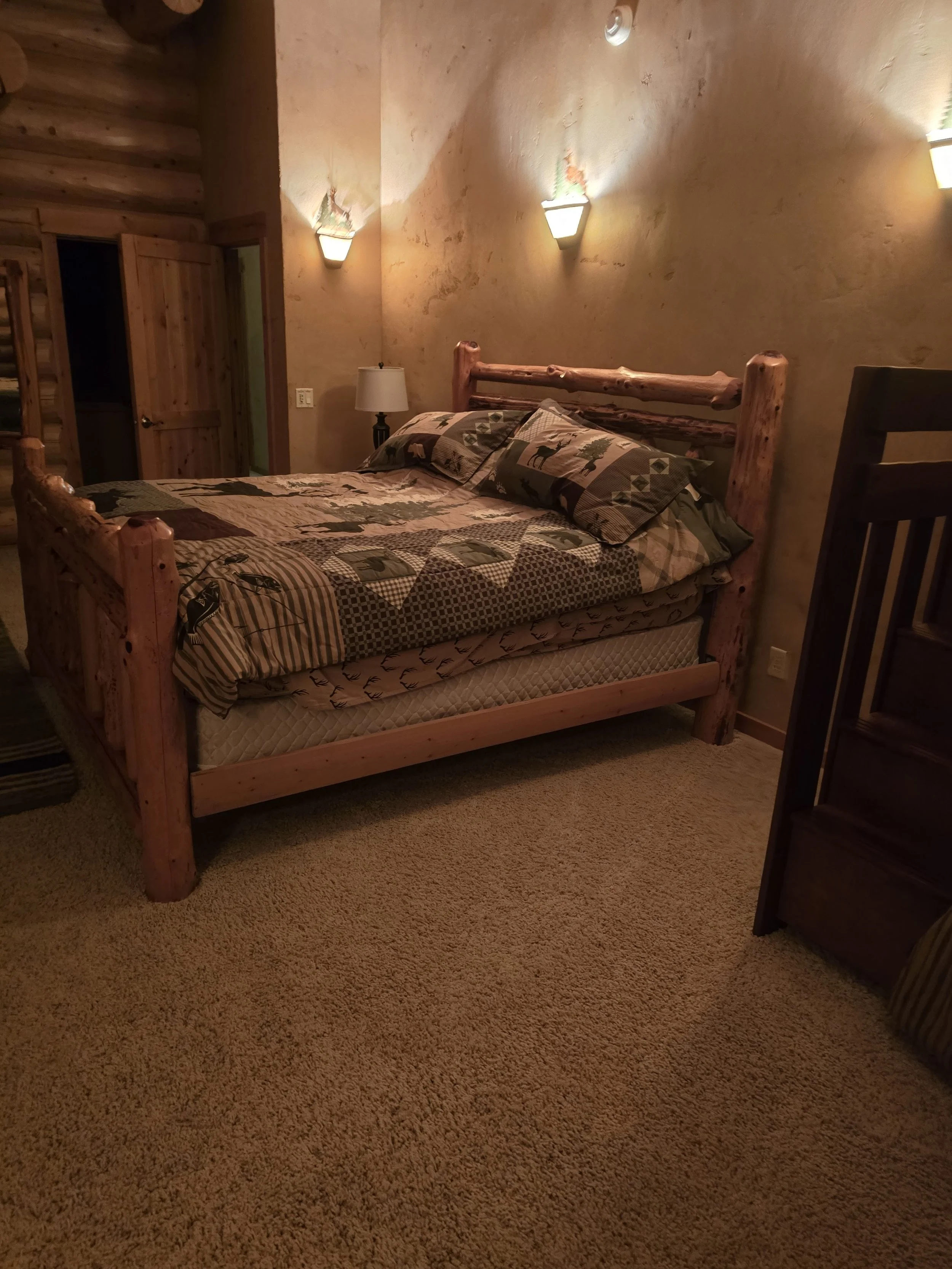 Rustic bedroom with a wooden bed frame and patchwork quilt, nightstand with lamp, carpeted floor, and warm lighting.