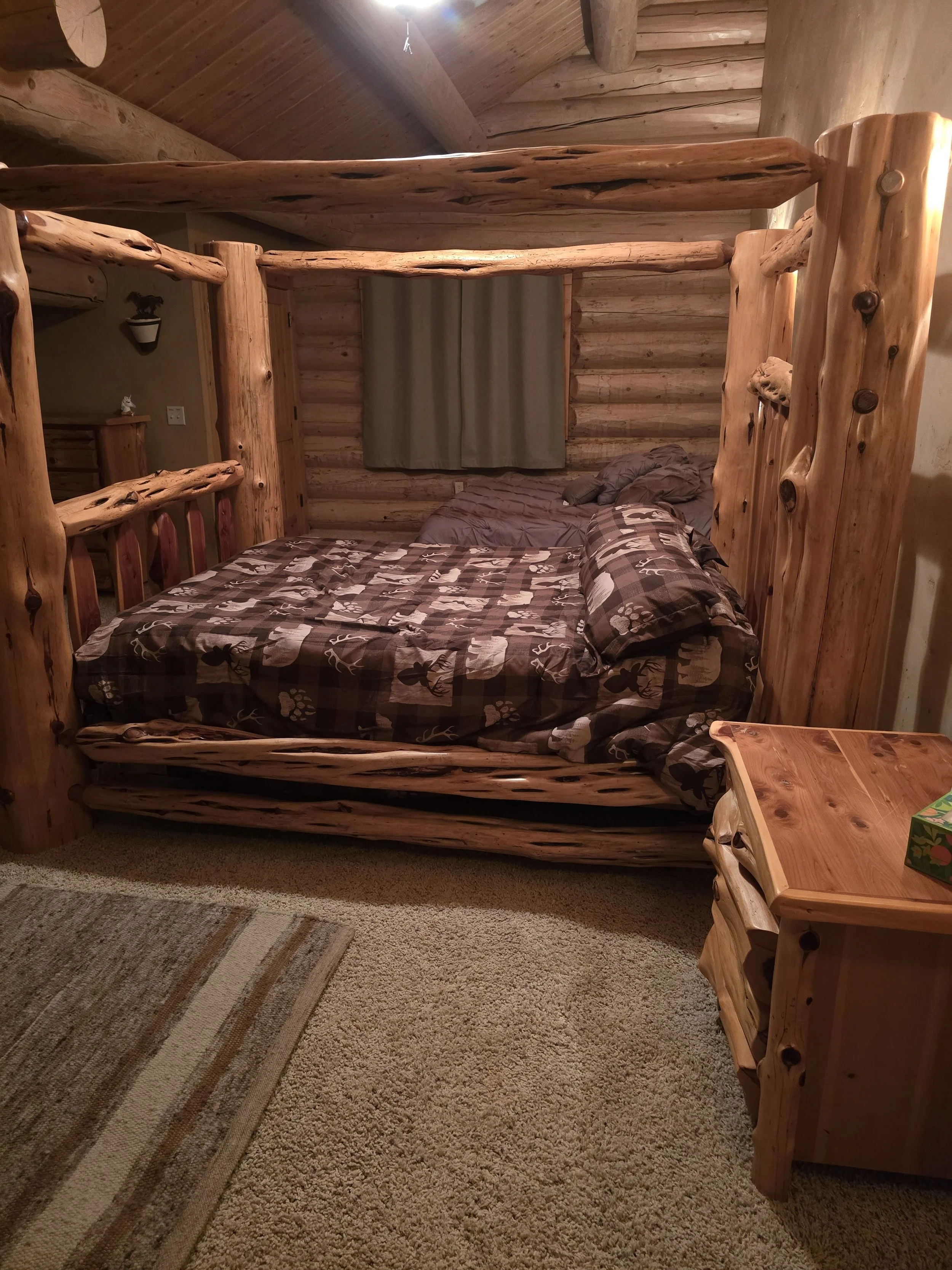 Rustic log cabin bedroom with a wooden bed frame, patterned bedding, wooden nightstand, wooden walls, and ceiling beams.