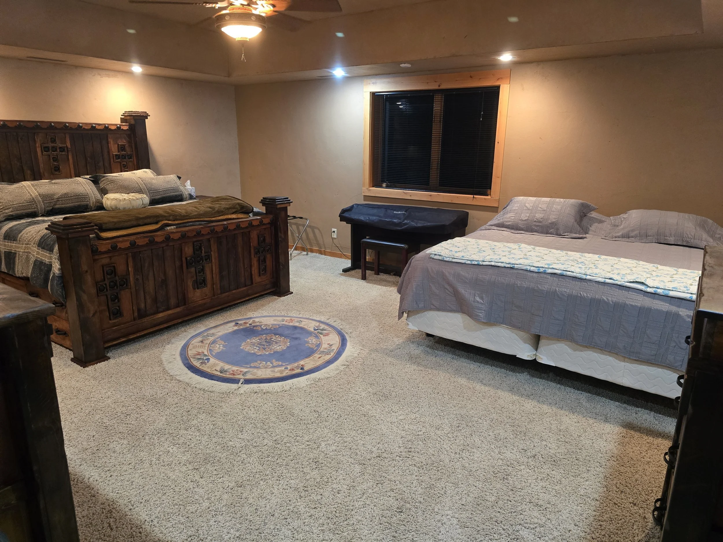 Bedroom with two beds, one large wooden bed and one smaller bed, carpeted floor, round rug, window with black blinds, ceiling fan.