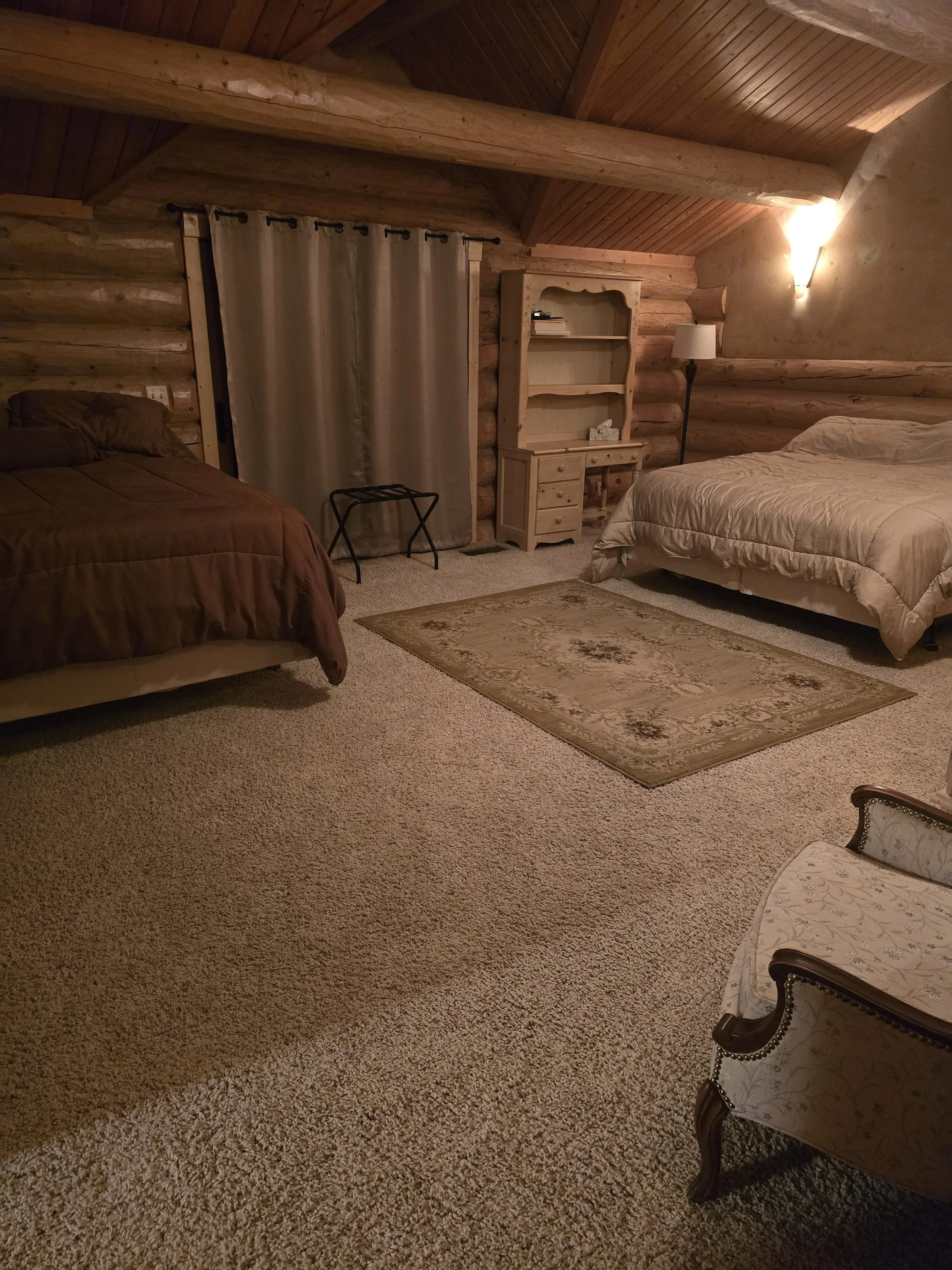 Cozy log cabin bedroom with exposed wooden beams and ceiling, featuring two beds covered in brown and beige bedding. The room includes a beige carpet, a wooden bookshelf, a small chair, and a curtain covering a window or closet.
