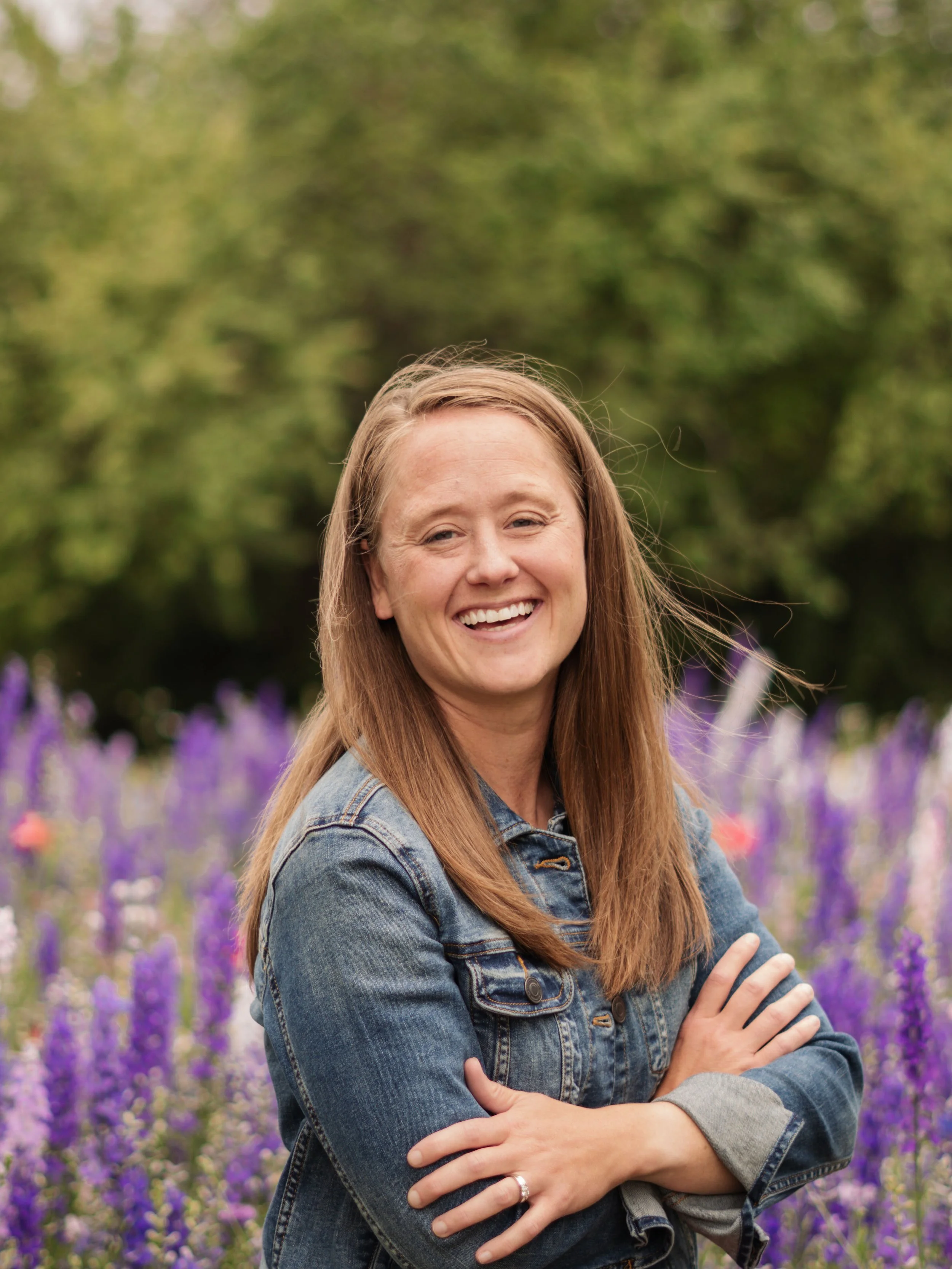 A woman with long red hair smiling and standing with arms crossed in a purple flower garden during daytime.
