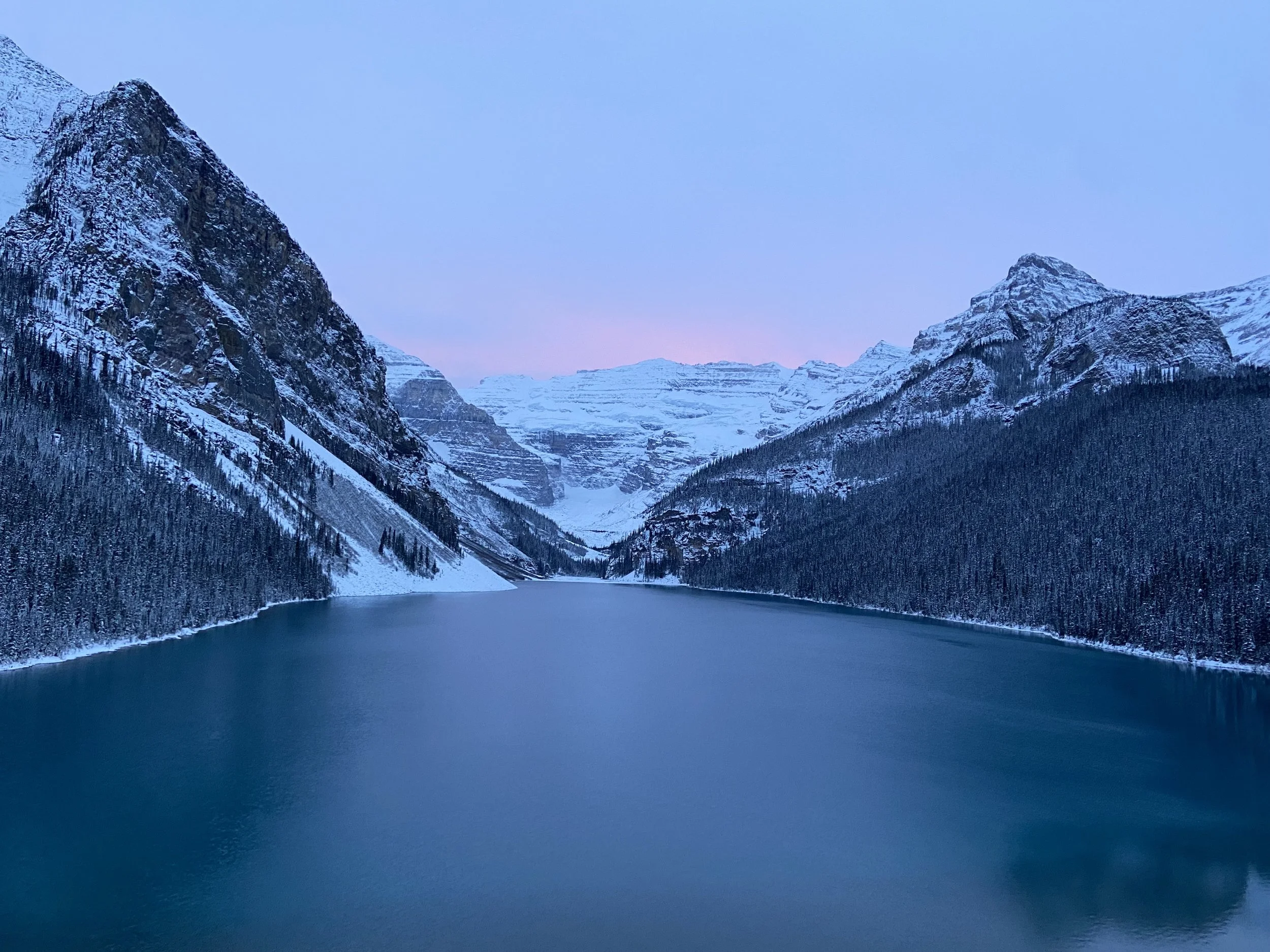 Snow-covered mountains surrounding a lake during sunset, with pink hues in the sky.