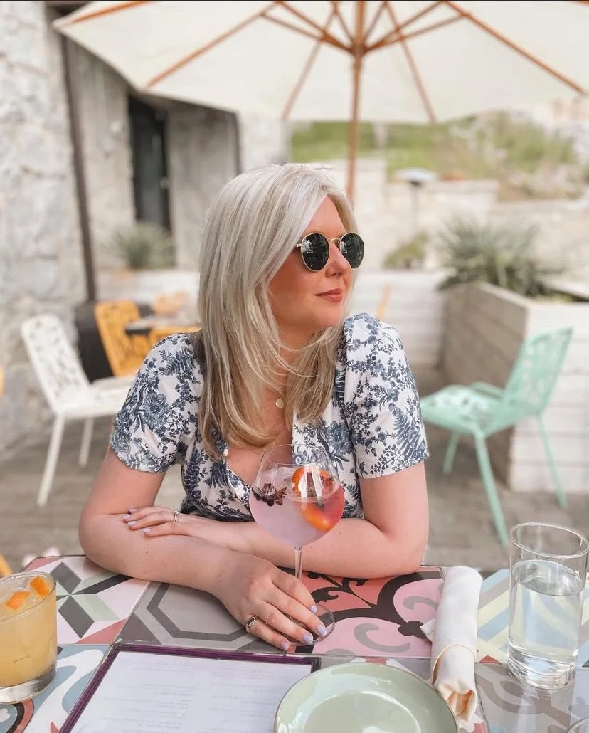 A person seated outdoors at a table under a large umbrella, holding a glass with a pink drink and citrus slices. They are wearing sunglasses and a floral outfit. The table has a decorative pattern and contains a menu, a rolled napkin, and additional drinks.