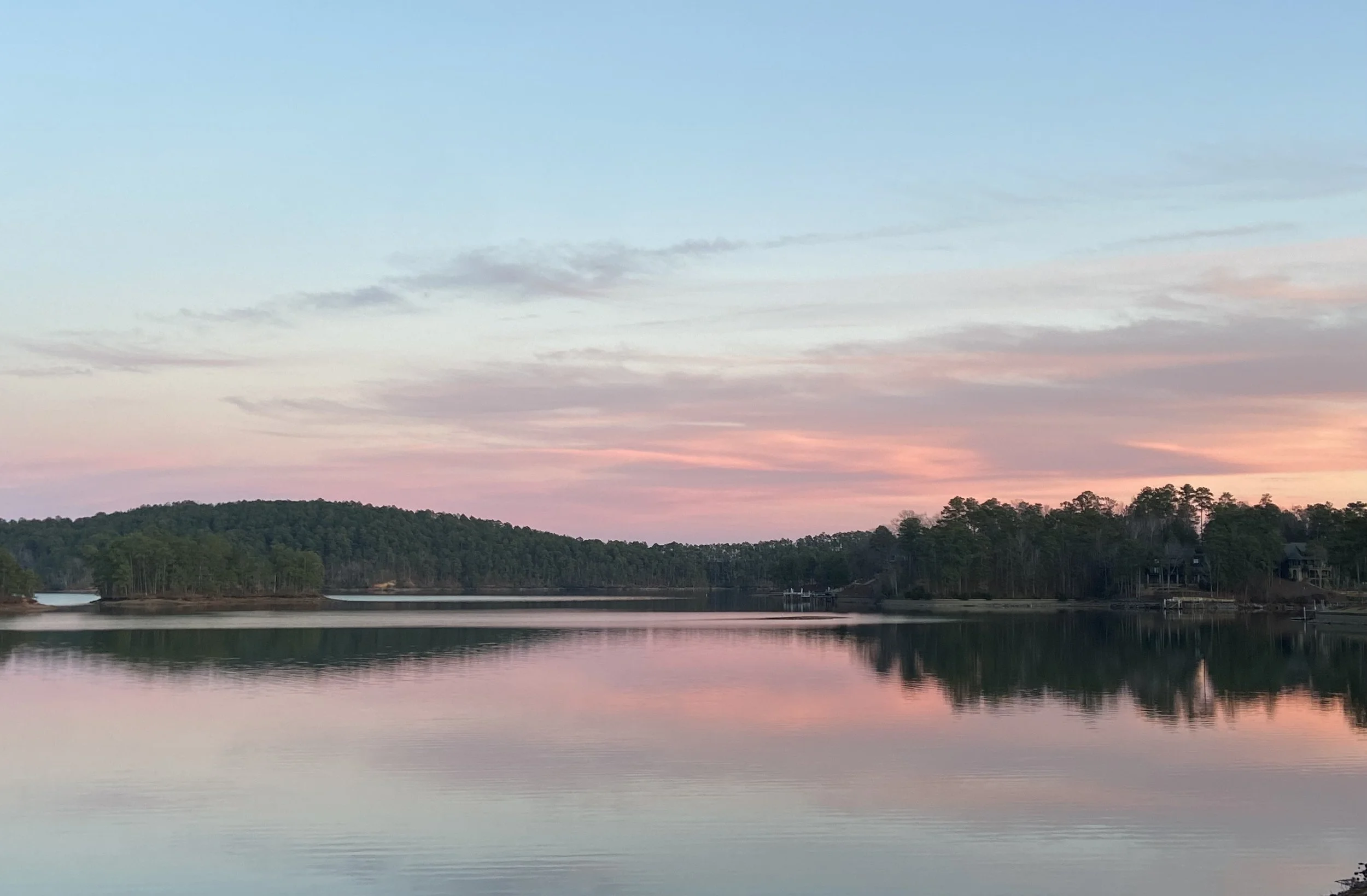 A serene lake at sunset with pink and blue skies, surrounded by a forested shoreline and gentle water reflections.
