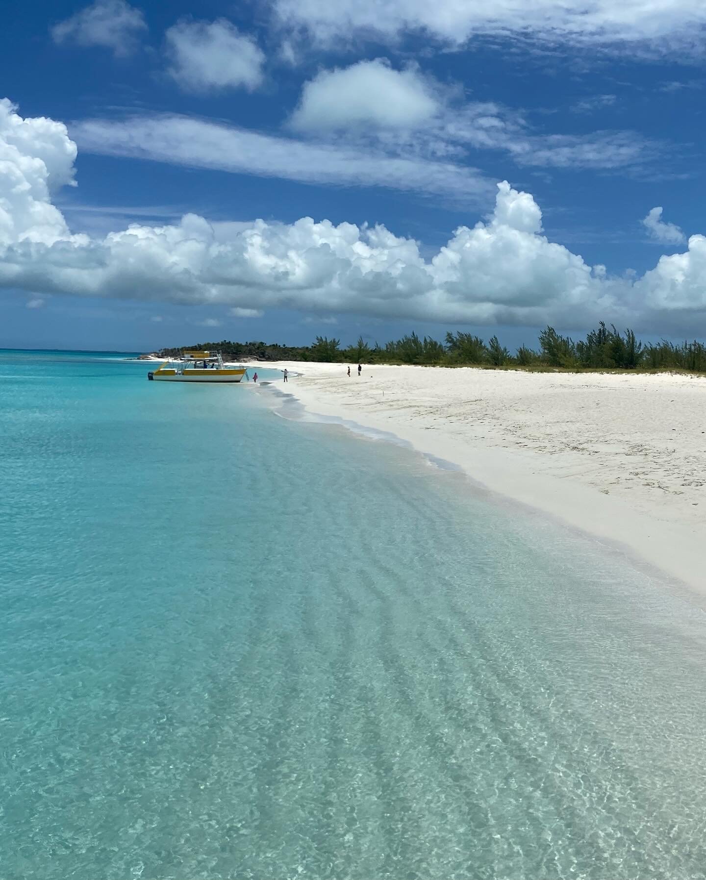 Tropical beach with clear turquoise water, white sand, and a blue sky with fluffy clouds. A yellow boat is docked on the shore.