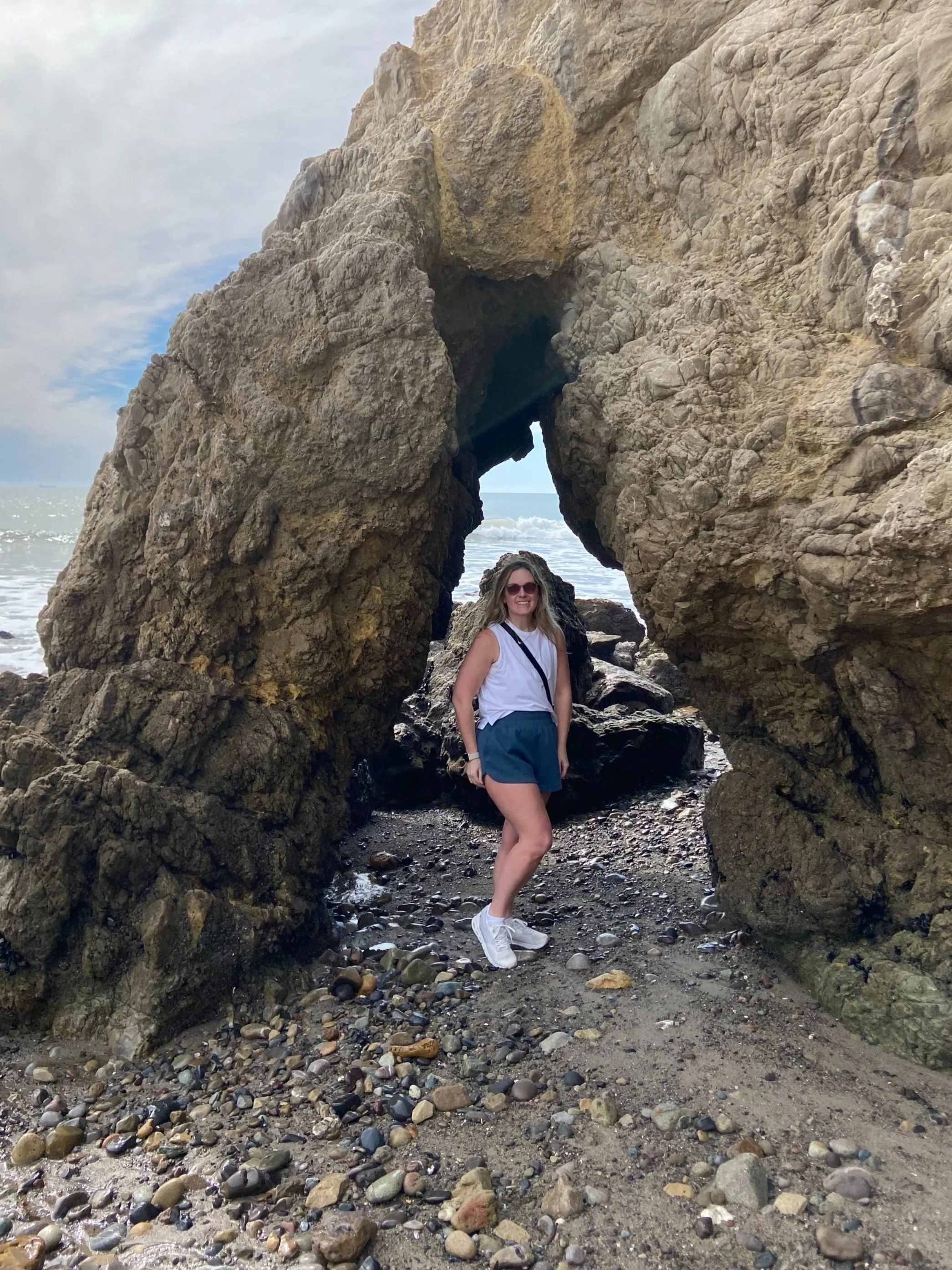 Person standing near a natural rock arch on a pebble beach with the sea and sky in the background.