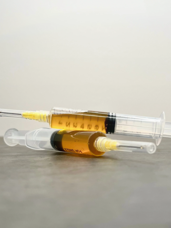 Two syringes filled with a dark amber liquid, lying on a flat surface against a plain background.