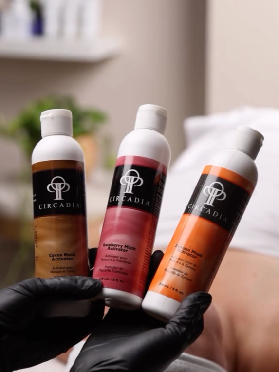 Person wearing black gloves holding three bottles of skincare products in a clinic or spa setting.
