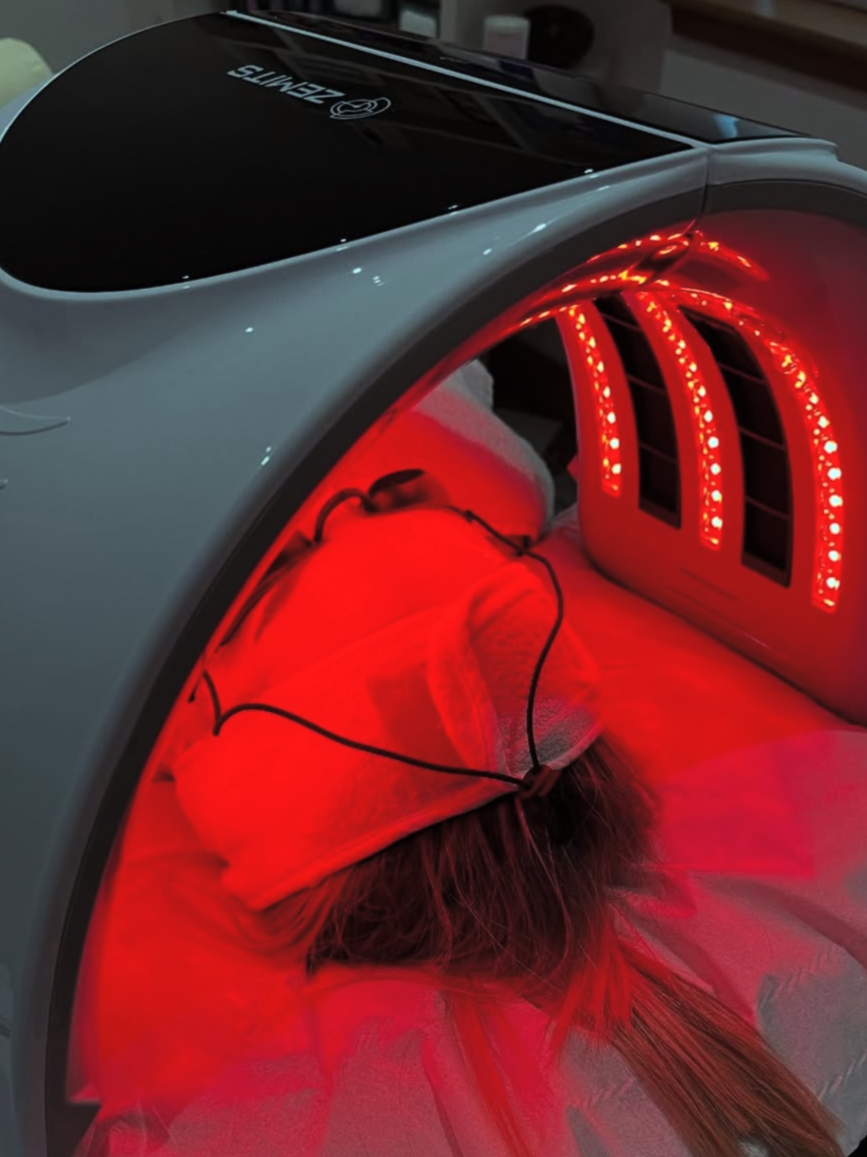 A person lying inside a red light therapy device, with their face covered by a pillow and wearing a face mask, under a heated bed or infrared sauna. The device has illuminated red LED lights and a curved design.