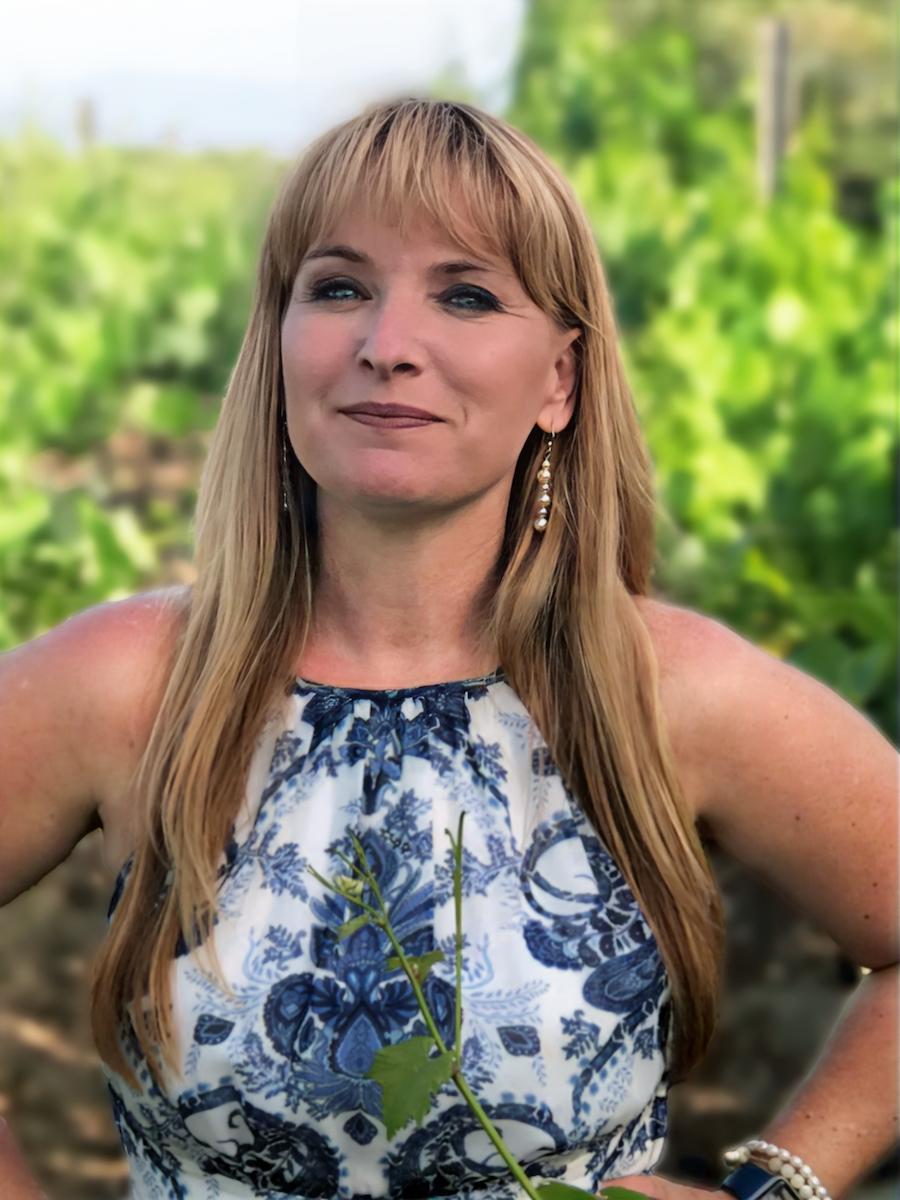 A woman with long blonde hair wearing a sleeveless blue and white floral dress, standing outdoors with a leafy green background.