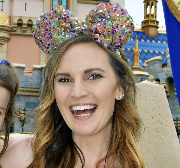 A woman smiling at an amusement park, wearing sparkly Minnie Mouse ears with colorful sequins, with castle-themed structures in the background.